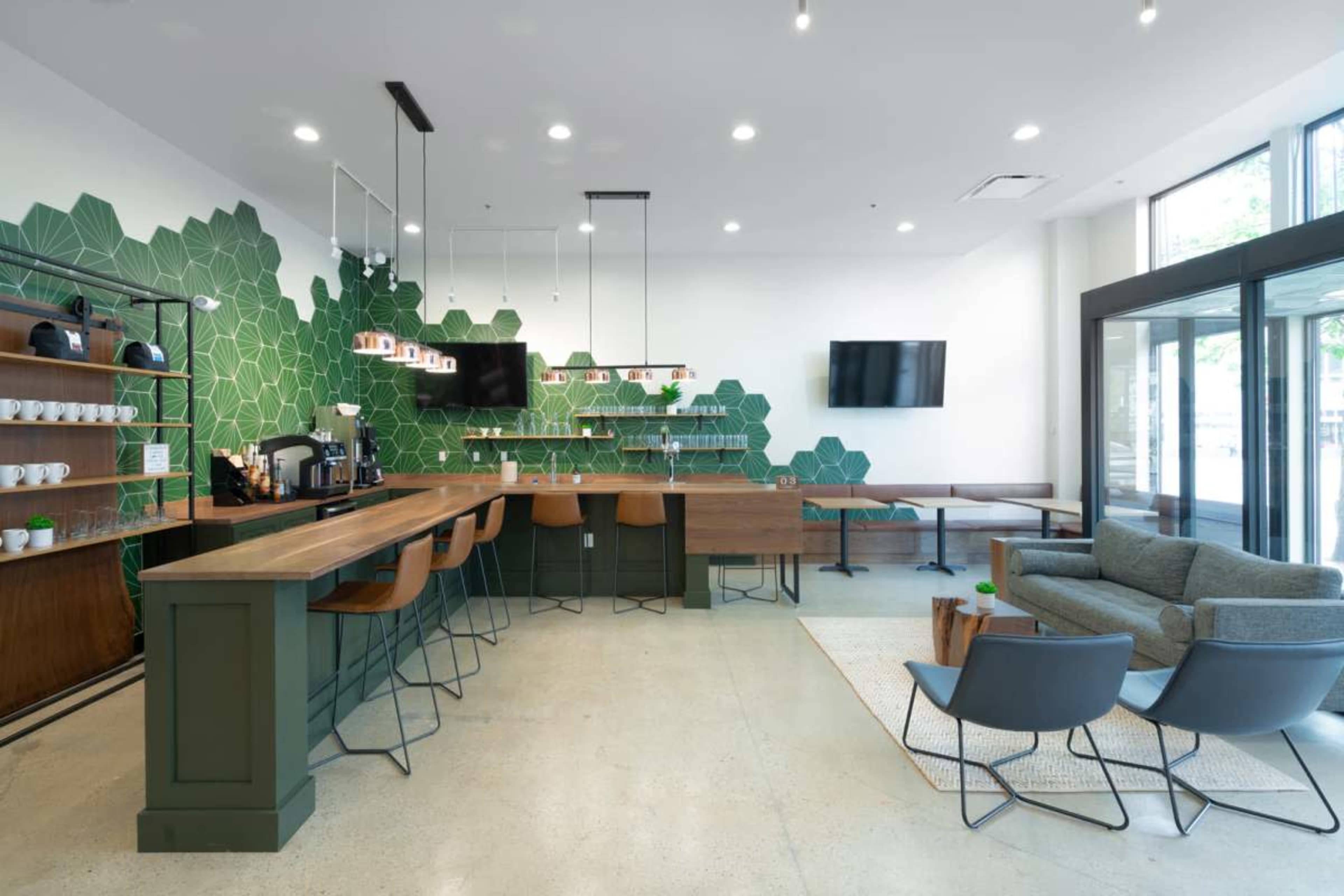 The image shows a modern café interior featuring a green geometric wall design, a wooden bar counter with stools, and comfortable seating areas.