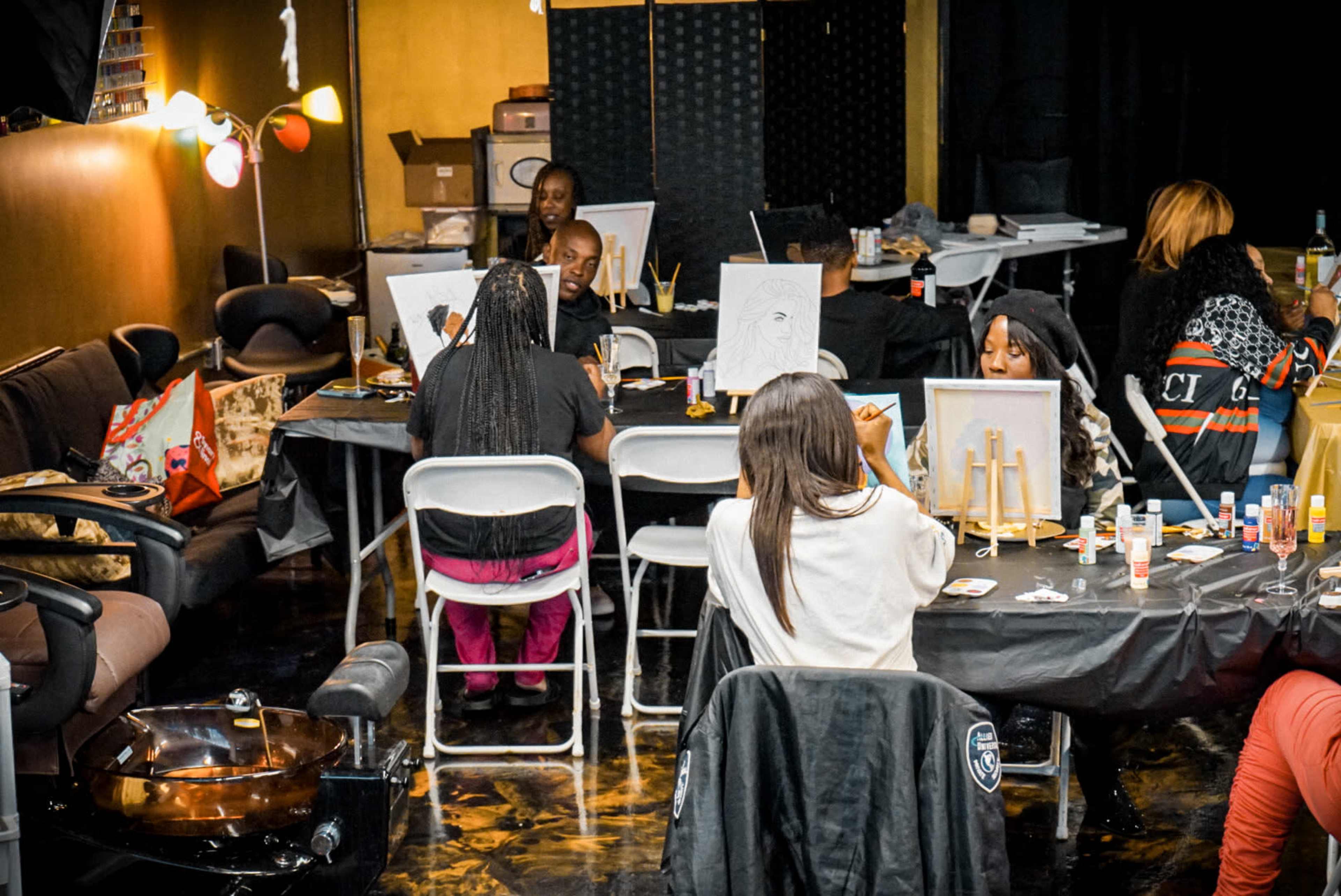 A group of people sits at tables in an art studio, painting on canvases while surrounded by various supplies and furniture.