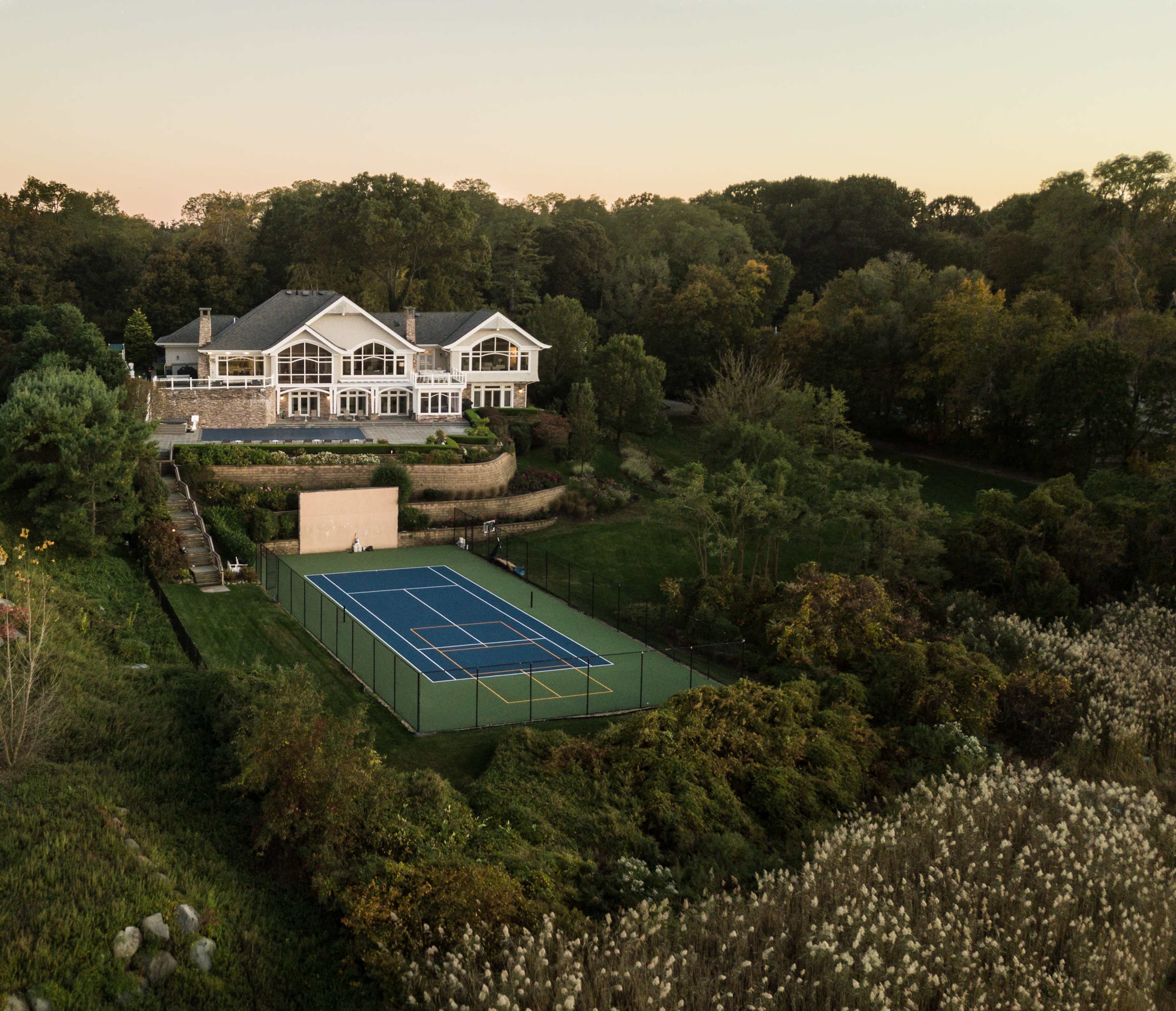 A luxurious house with multiple balconies overlooks a green tennis court surrounded by landscaping at sunset.