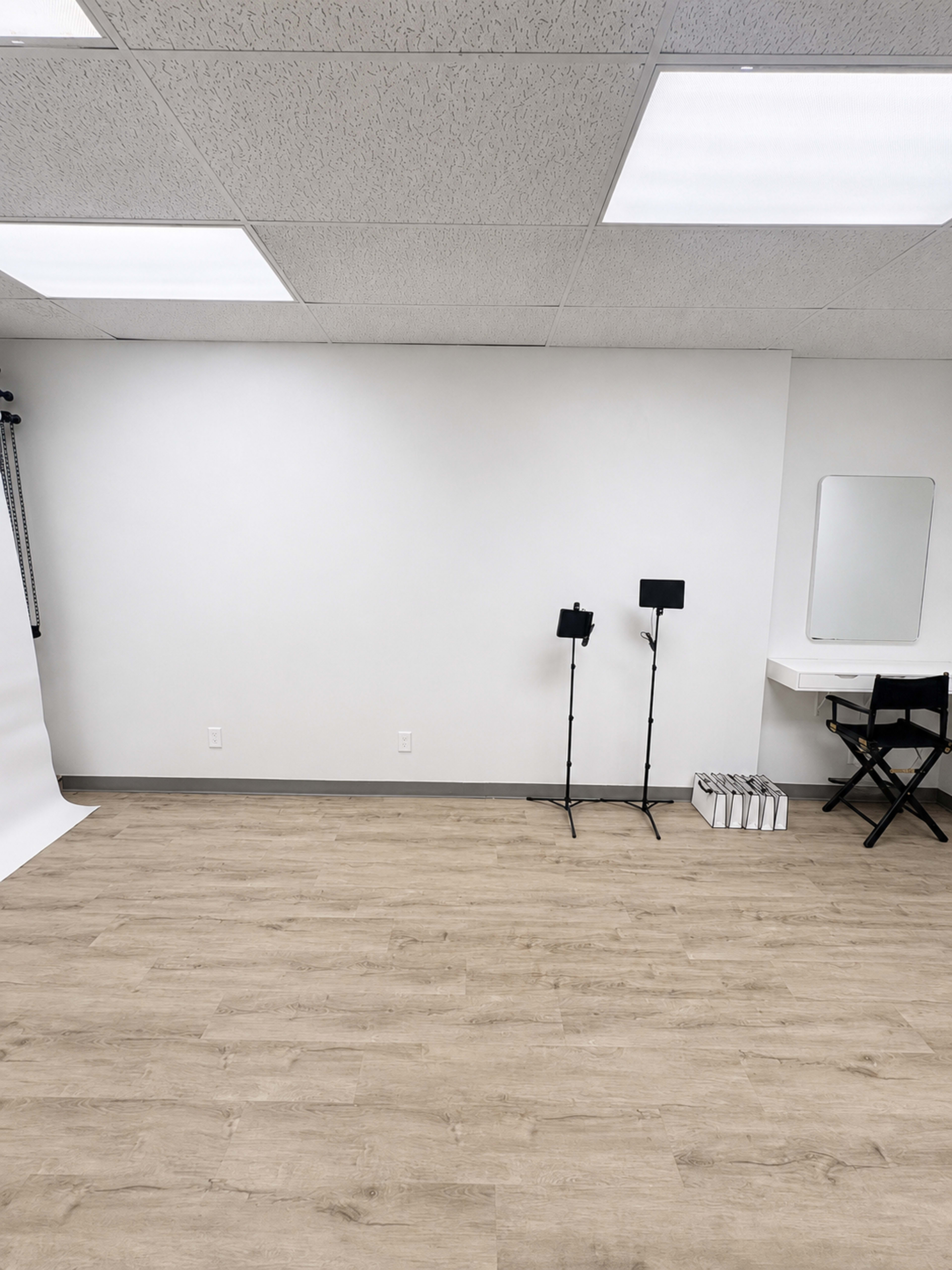 The image shows an empty room with light wood flooring, a white wall, and two equipment stands next to a dressing table and a director's chair.