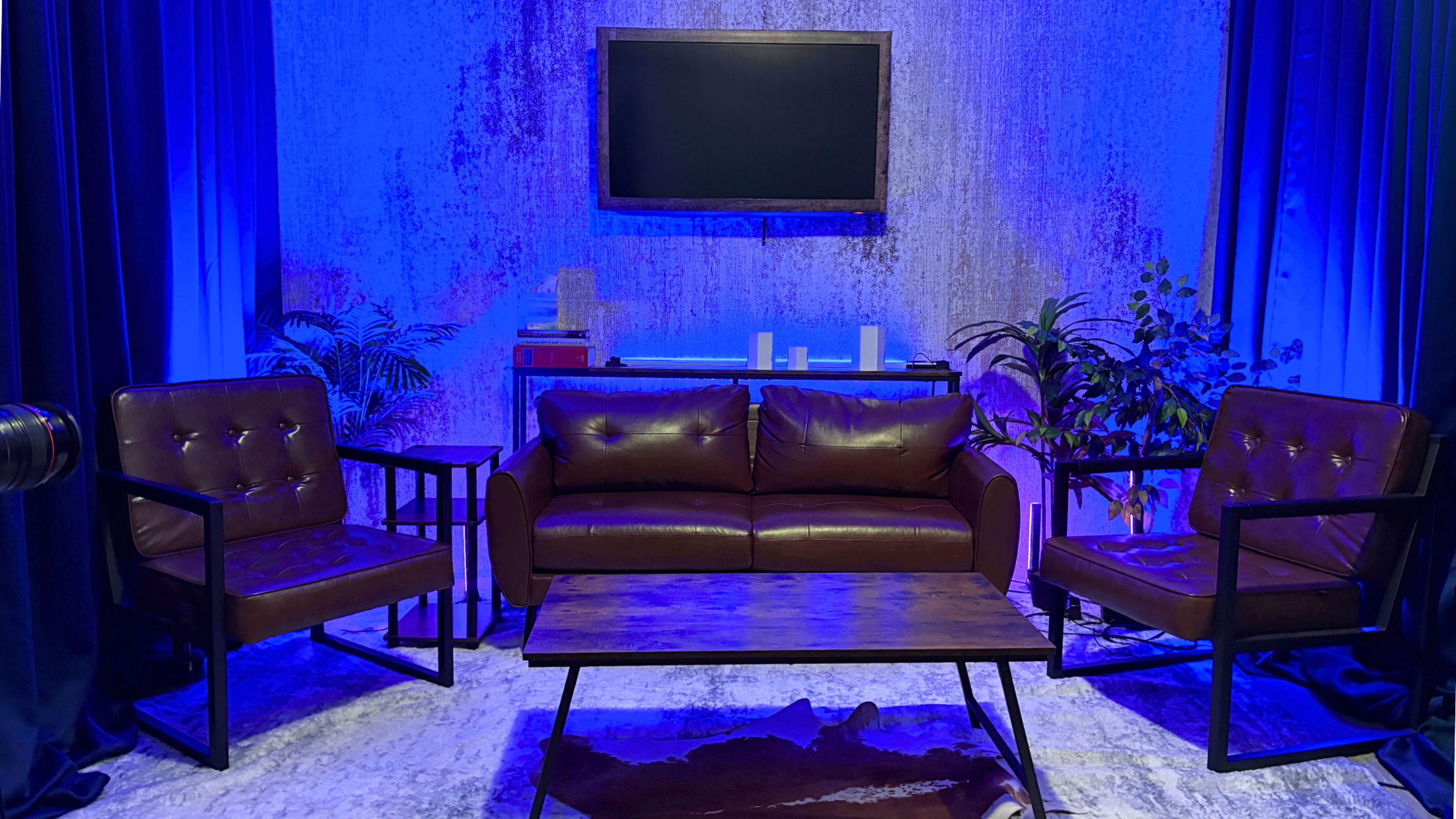 The image shows a modern living room setup with a brown leather sofa, two matching armchairs, a wooden coffee table, and a television mounted on a textured wall, all illuminated by blue lighting.