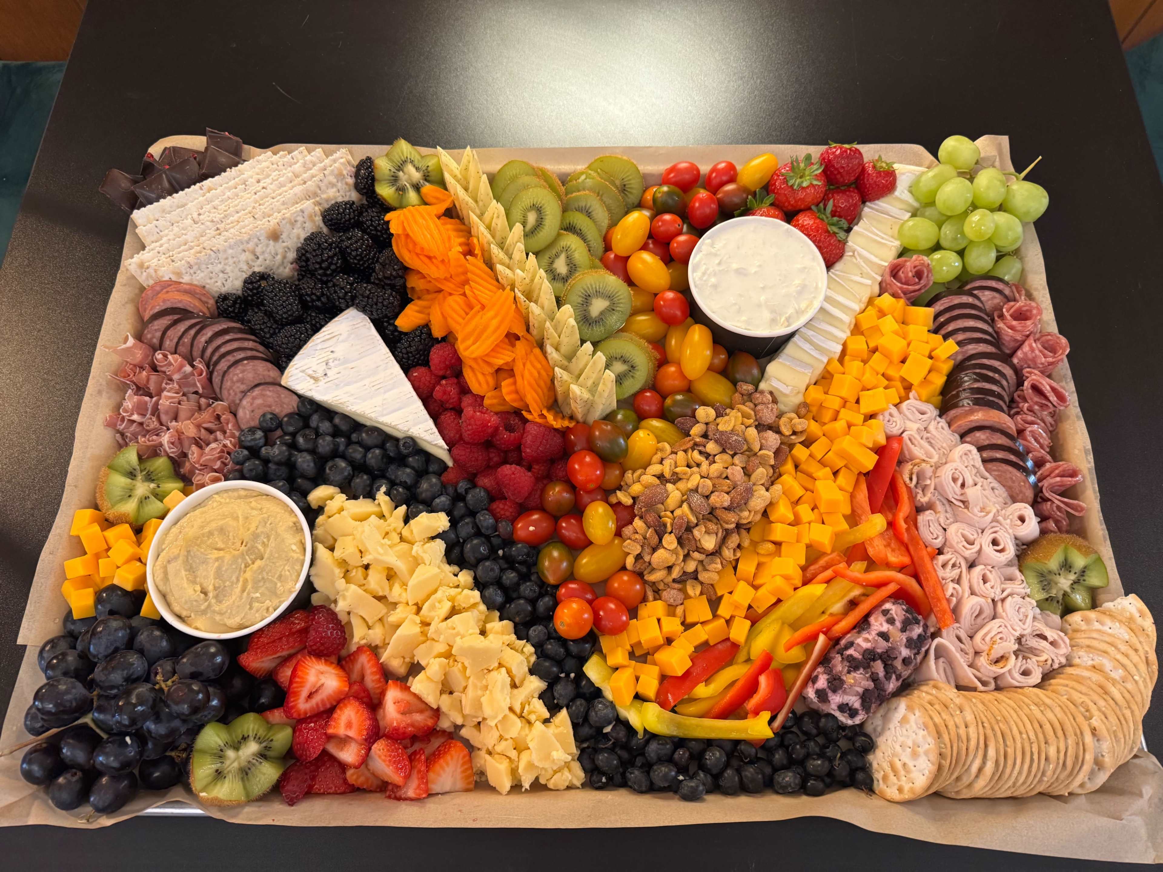 The image shows a large charcuterie board arranged with a variety of meats, cheeses, fruits, nuts, and dips, all neatly organized on a parchment-lined tray.