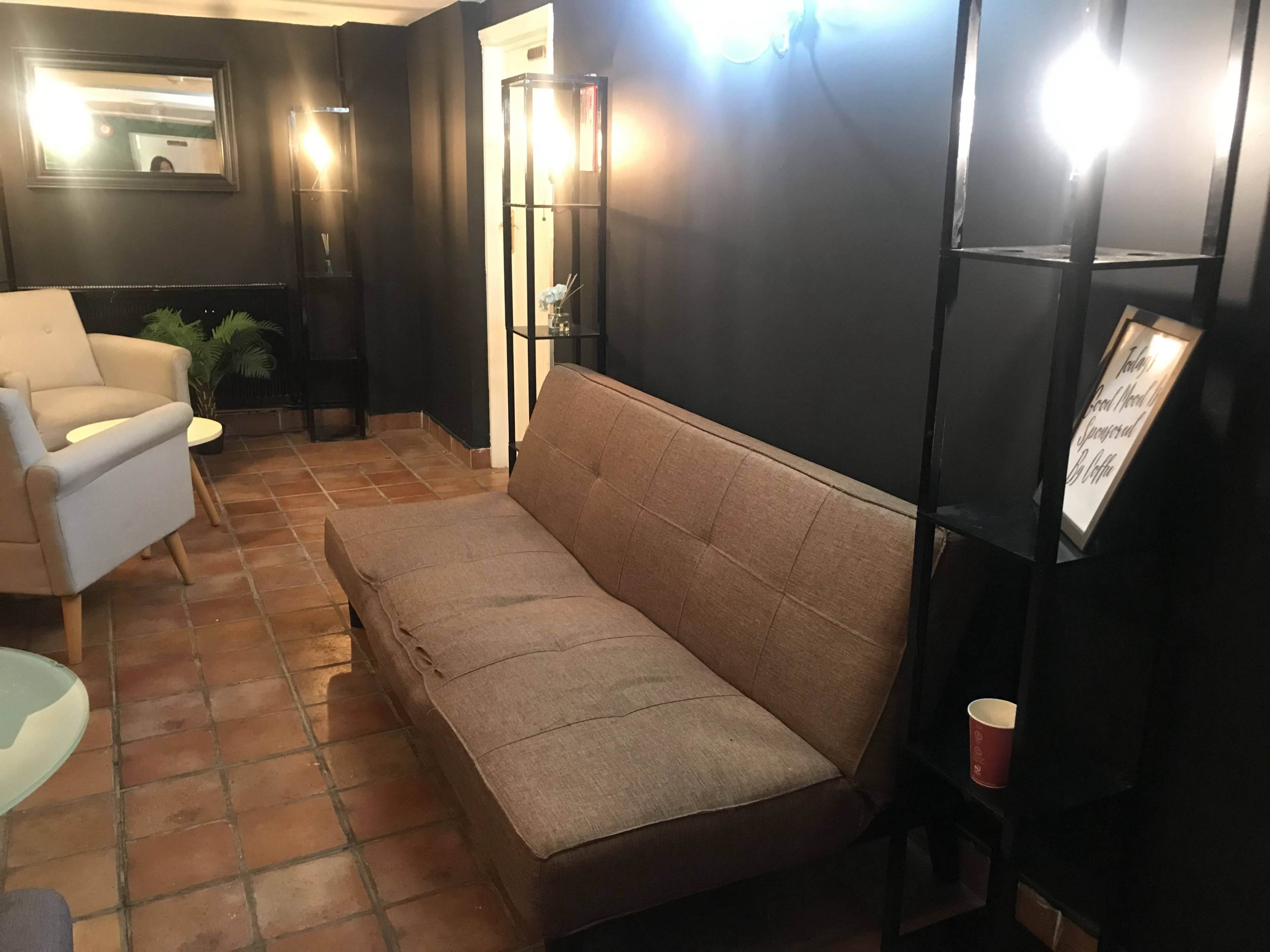 A cozy lounge area featuring a brown sofa, two light-colored armchairs, a shelf unit, and a glass table on tiled flooring.
