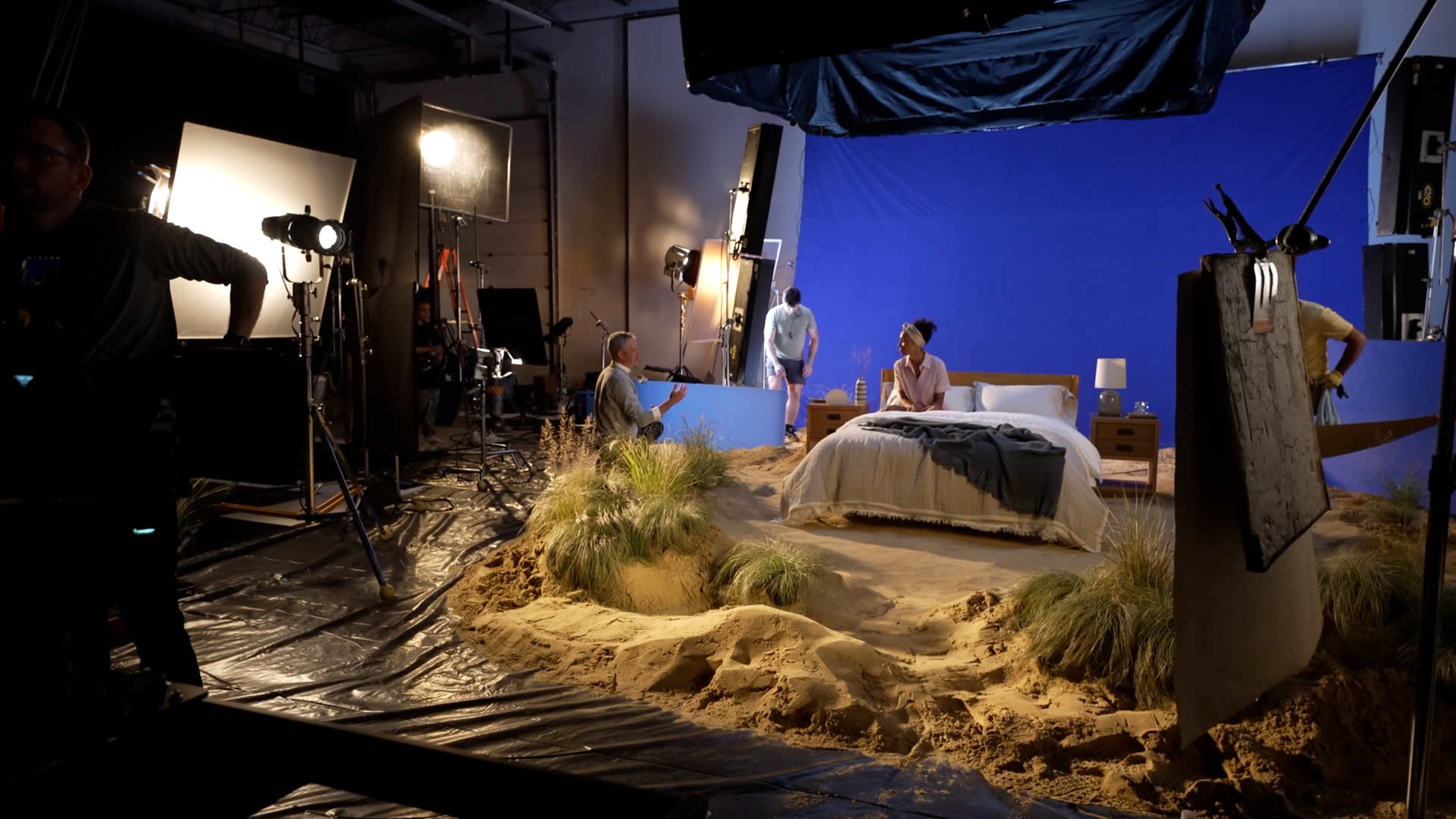 The scene depicts a film set with a bed in a sandy environment, surrounded by crew members and studio lighting equipment against a blue backdrop.