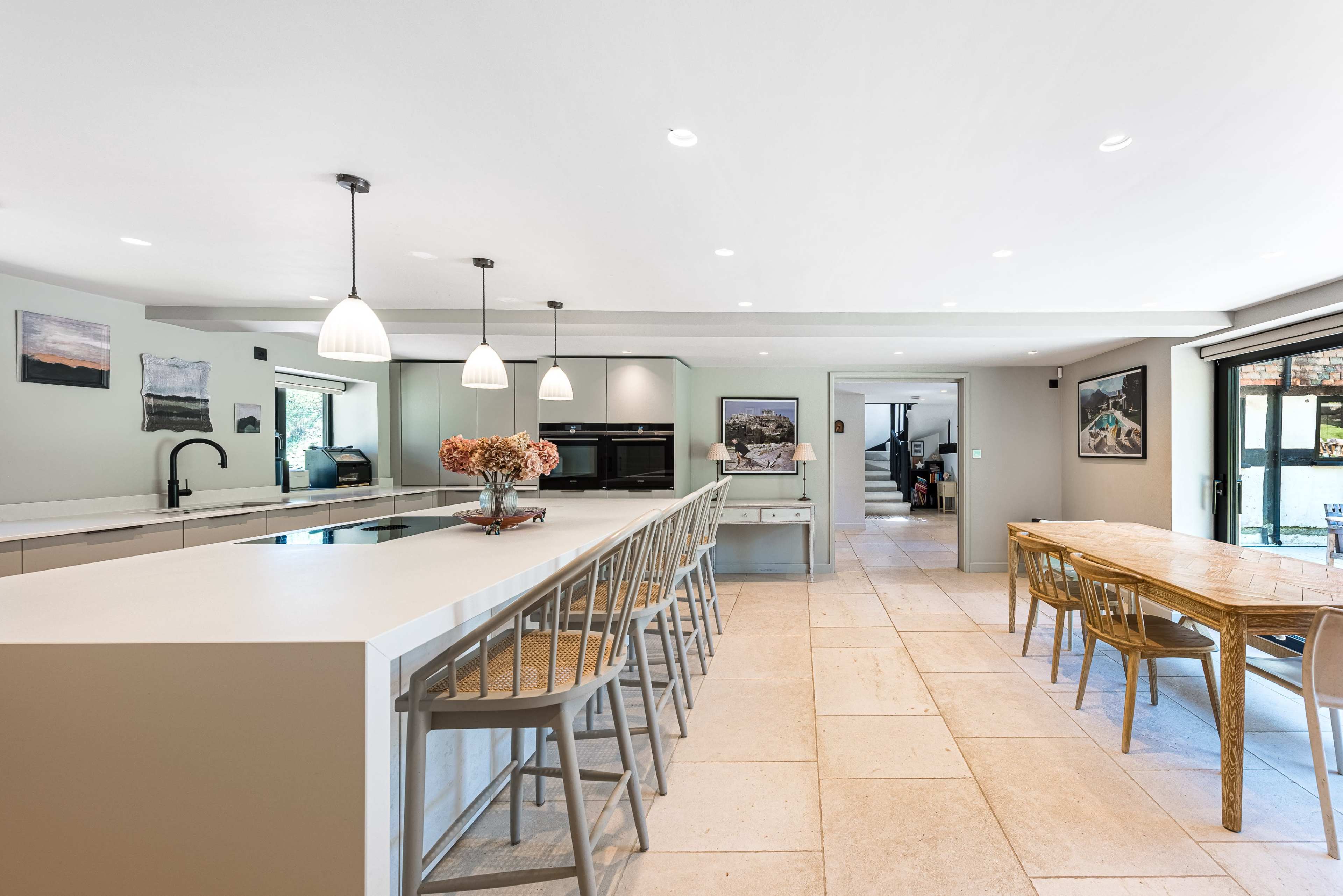 The image shows a modern kitchen with an island bar featuring seating, light fixtures overhead, and a dining area with a large table.