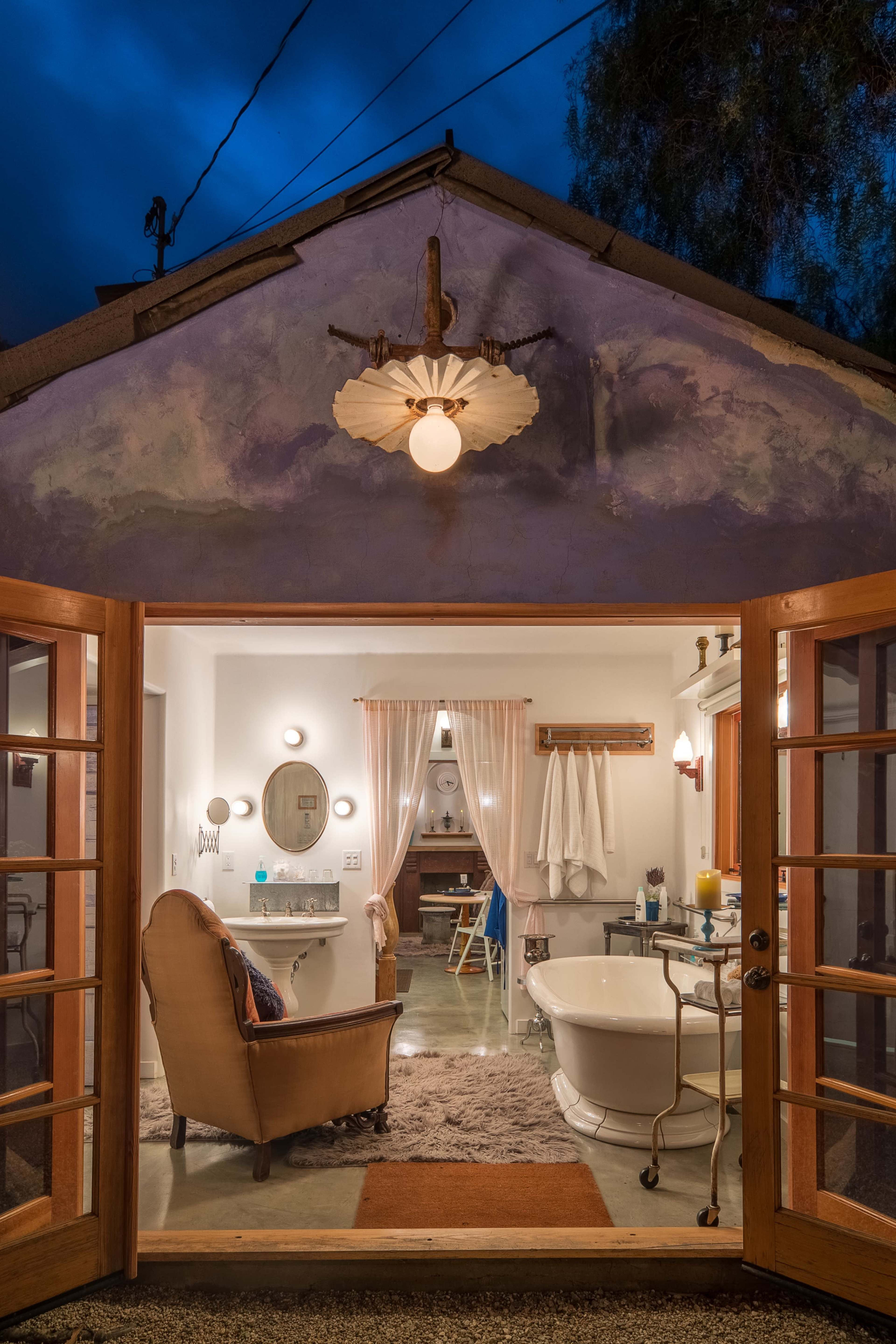A cozy bathroom with a freestanding bathtub, a vintage chair, and dual French doors opening to an evening sky.