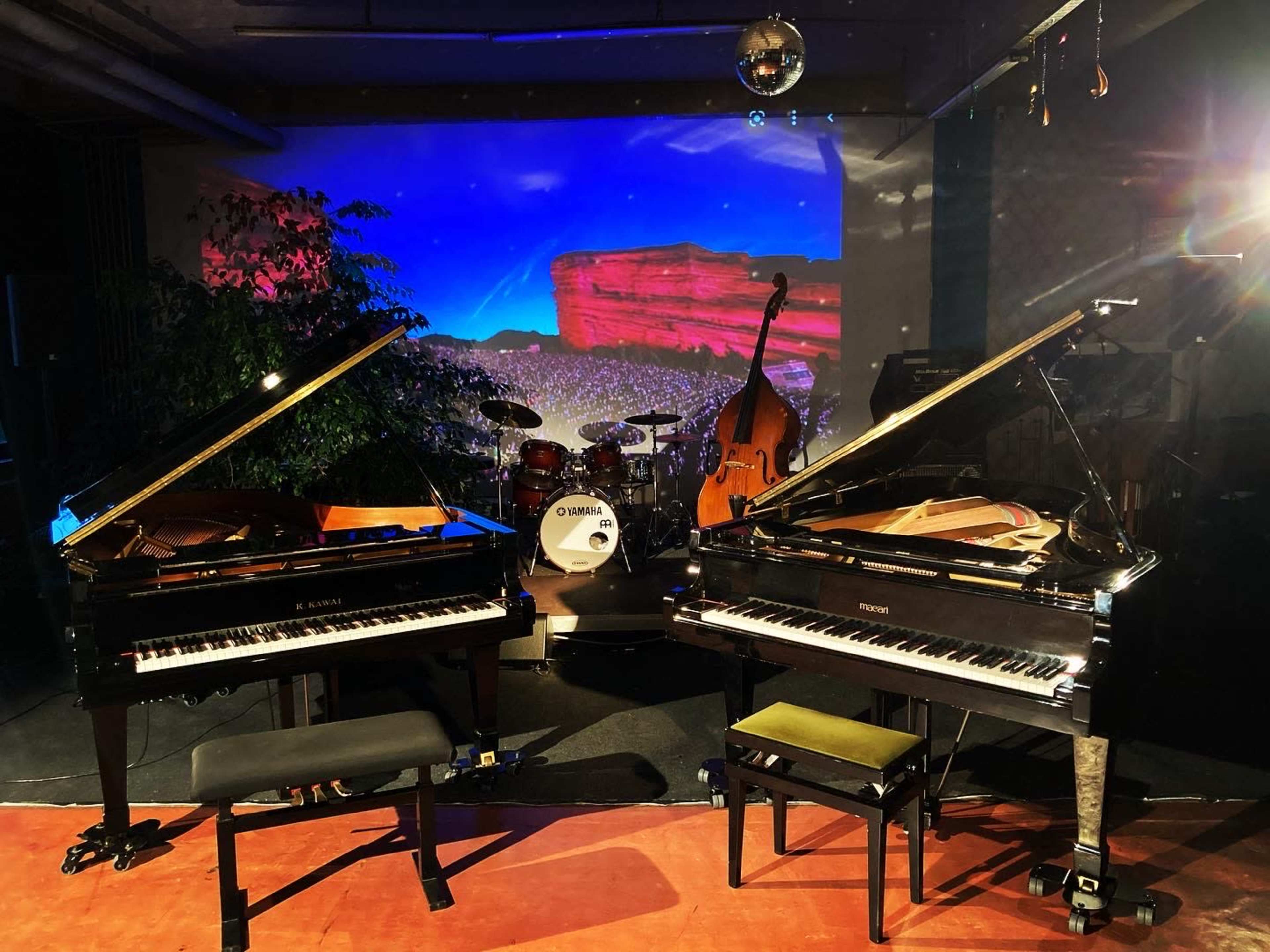 Two grand pianos are positioned on a stage with a backdrop of a scenic projection and a small band setup, including a drum set and double bass.