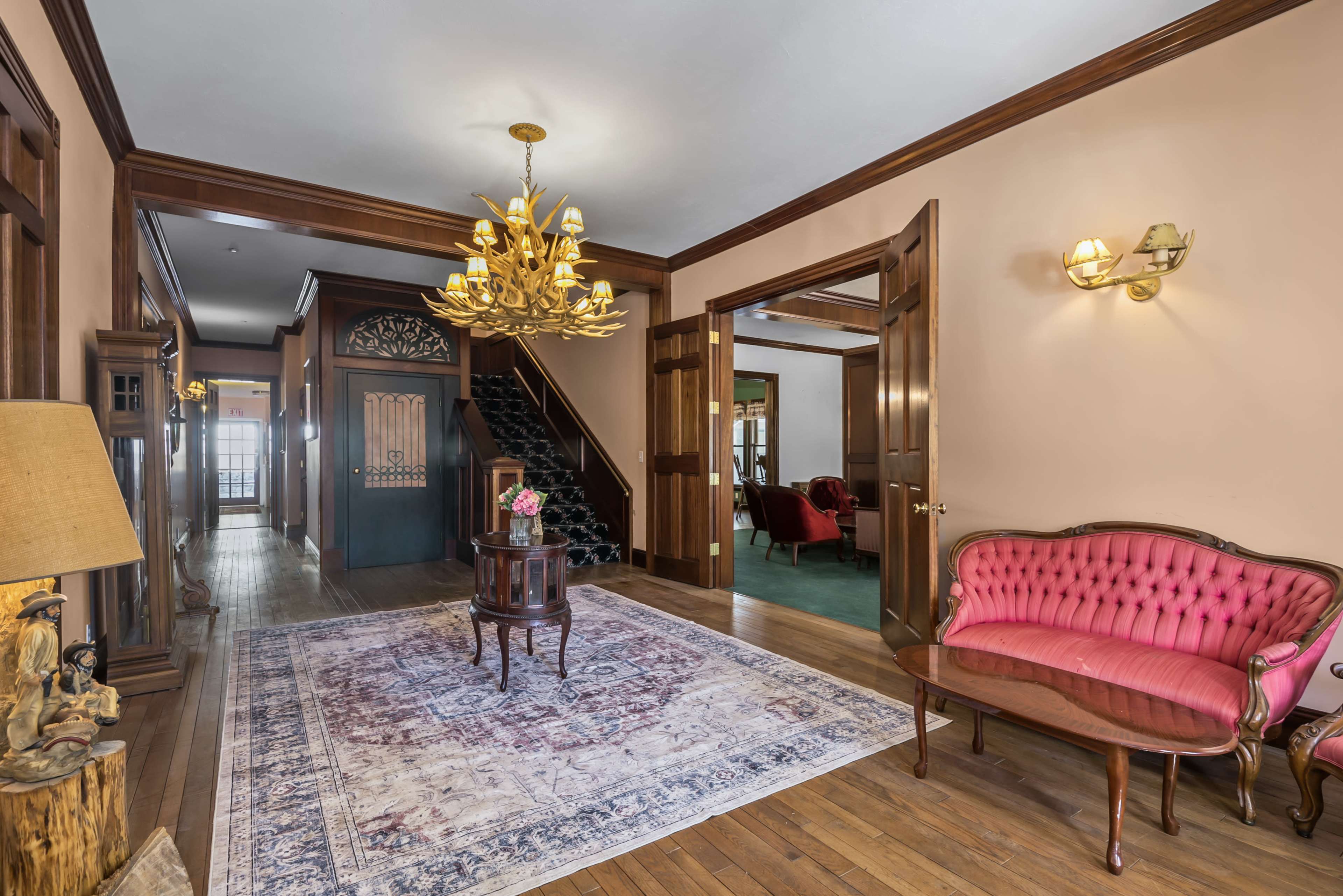 A vintage-style entryway featuring a pink upholstered settee, a wooden table, and a staircase leading to an upper level, with ornate lighting fixtures and a decorative rug on wooden flooring.