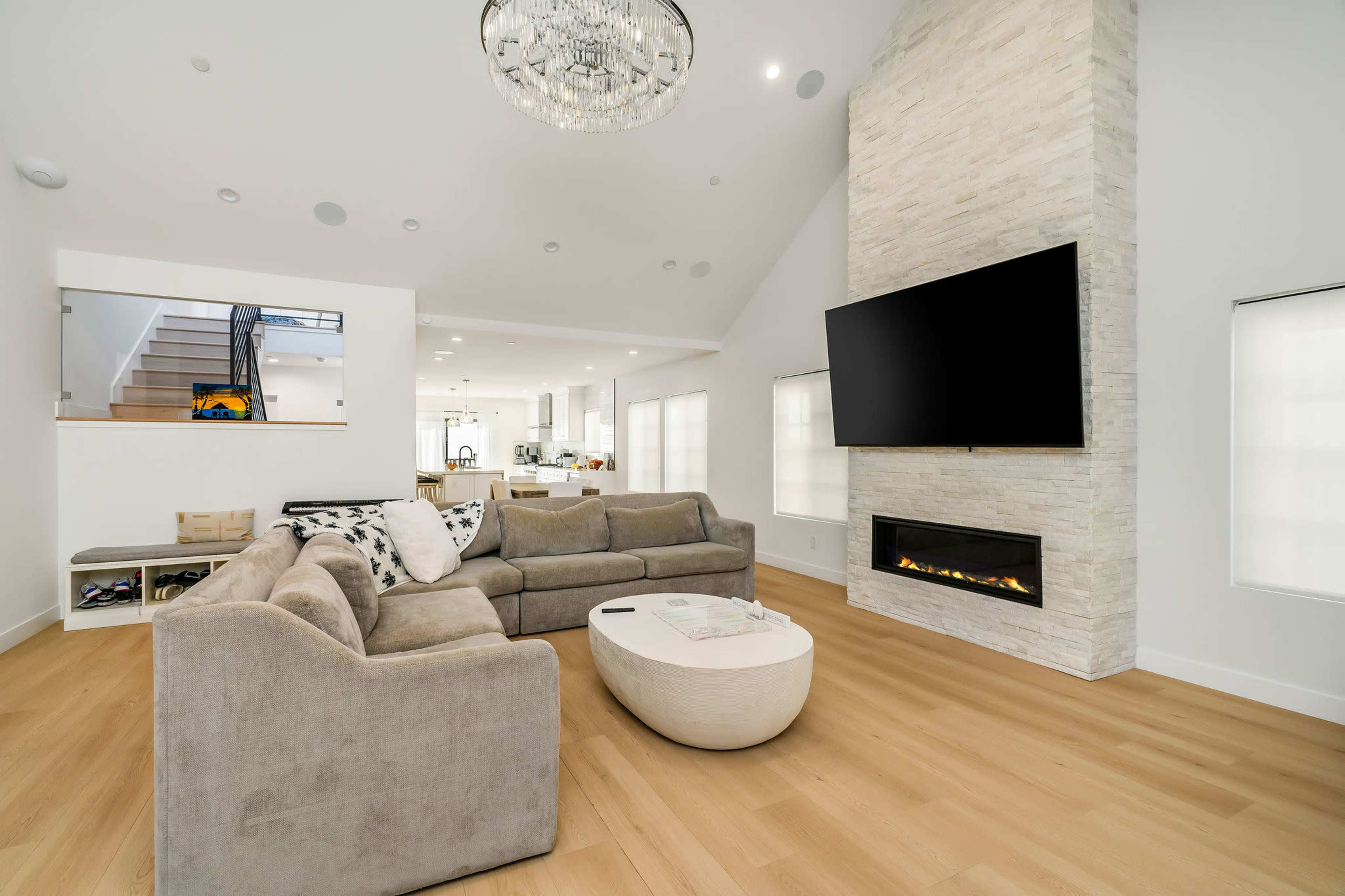 The image shows a modern living room featuring a large sectional sofa, a round coffee table, a wall-mounted TV above a fireplace, and a staircase in the background.