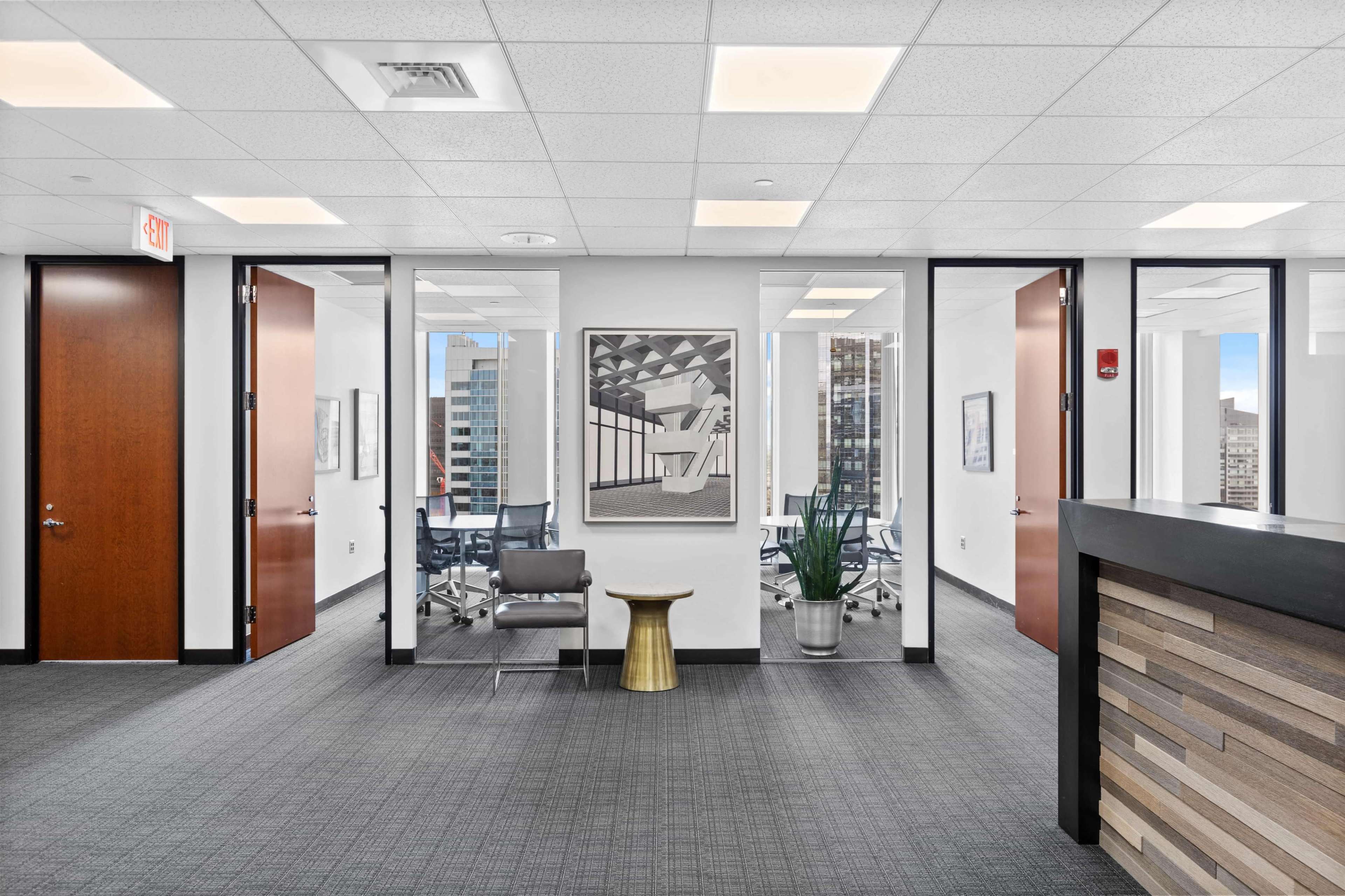 The image shows a modern office space featuring a reception area with a wooden desk, a decorative plant, and glass-walled meeting rooms in the background.