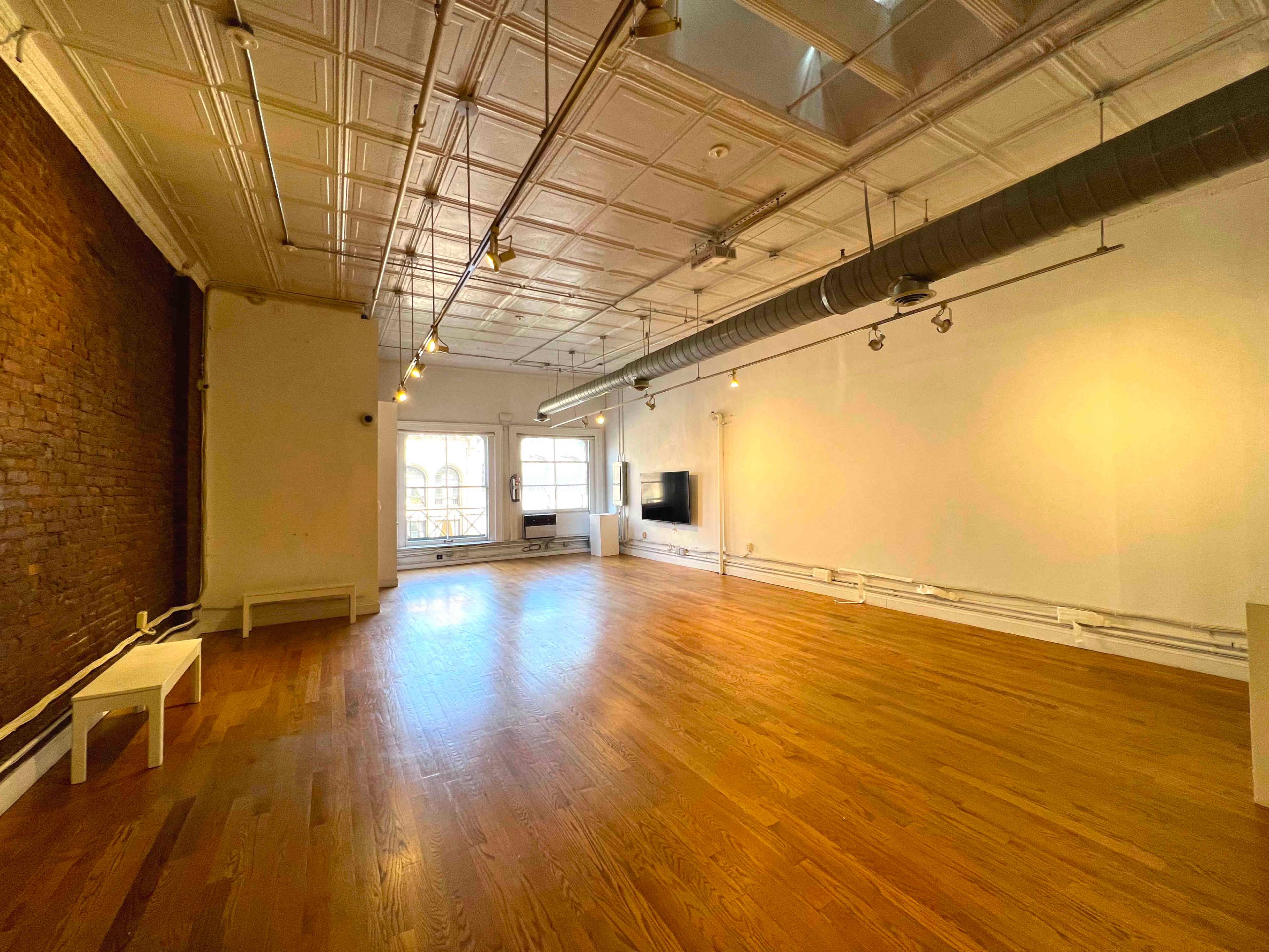 An empty, well-lit room with wooden floors, exposed brick walls, and a large window, featuring a sectional area for seating and a wall-mounted television.
