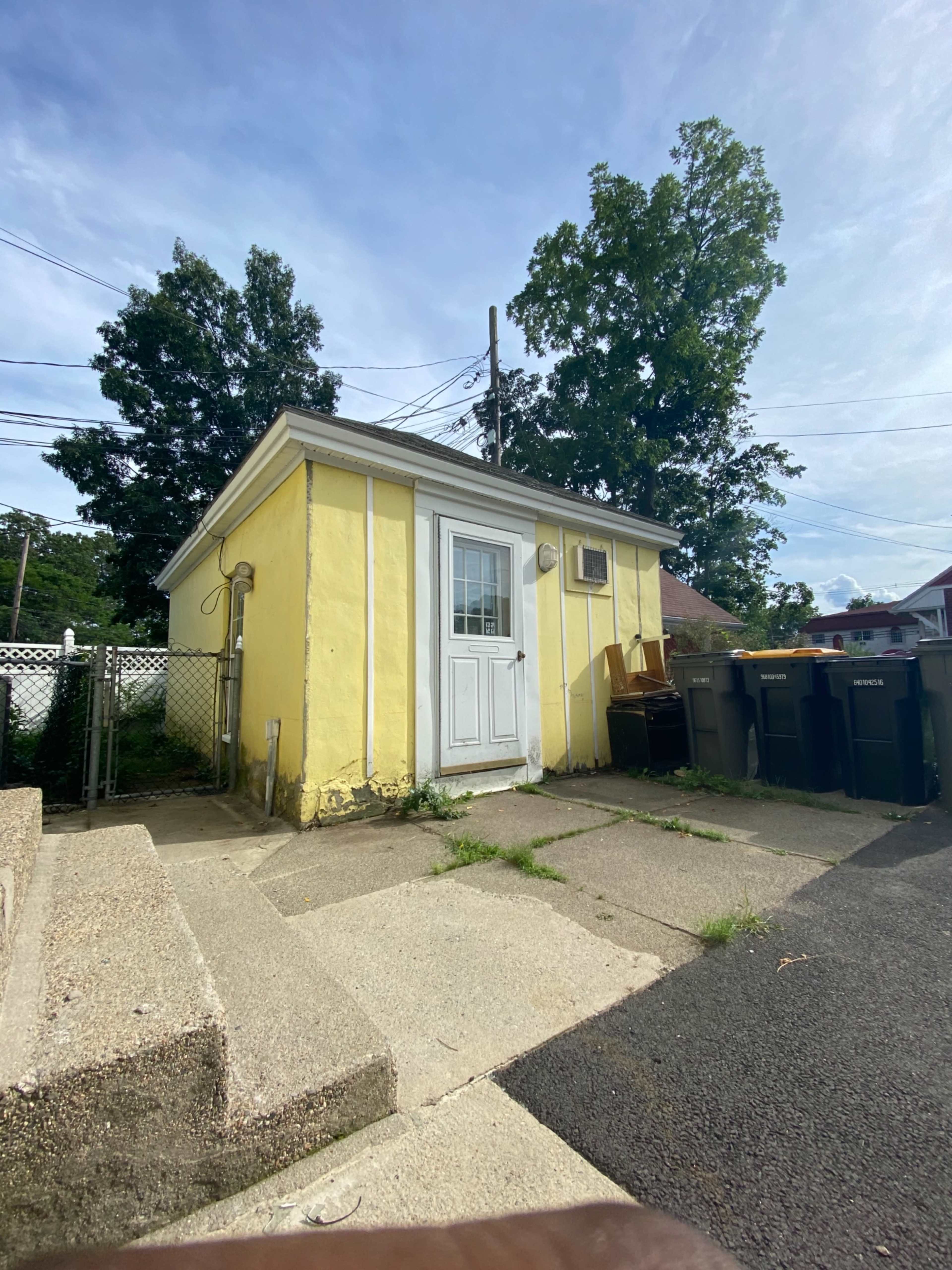 A small yellow shed with a white door and an air conditioning unit sits next to a pathway and several trash bins.