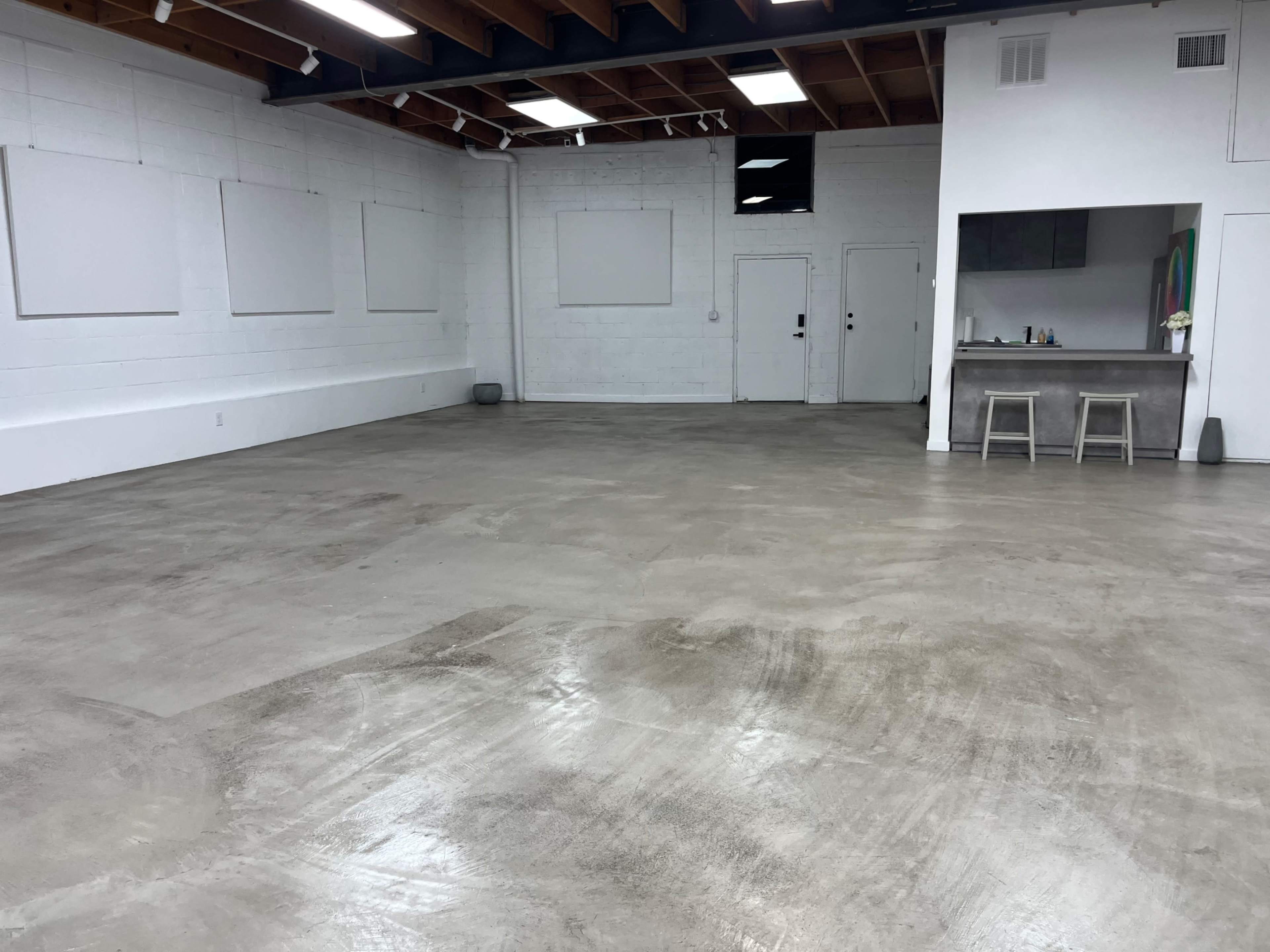 The image shows a spacious, empty room with concrete flooring and a small kitchenette area in one corner.