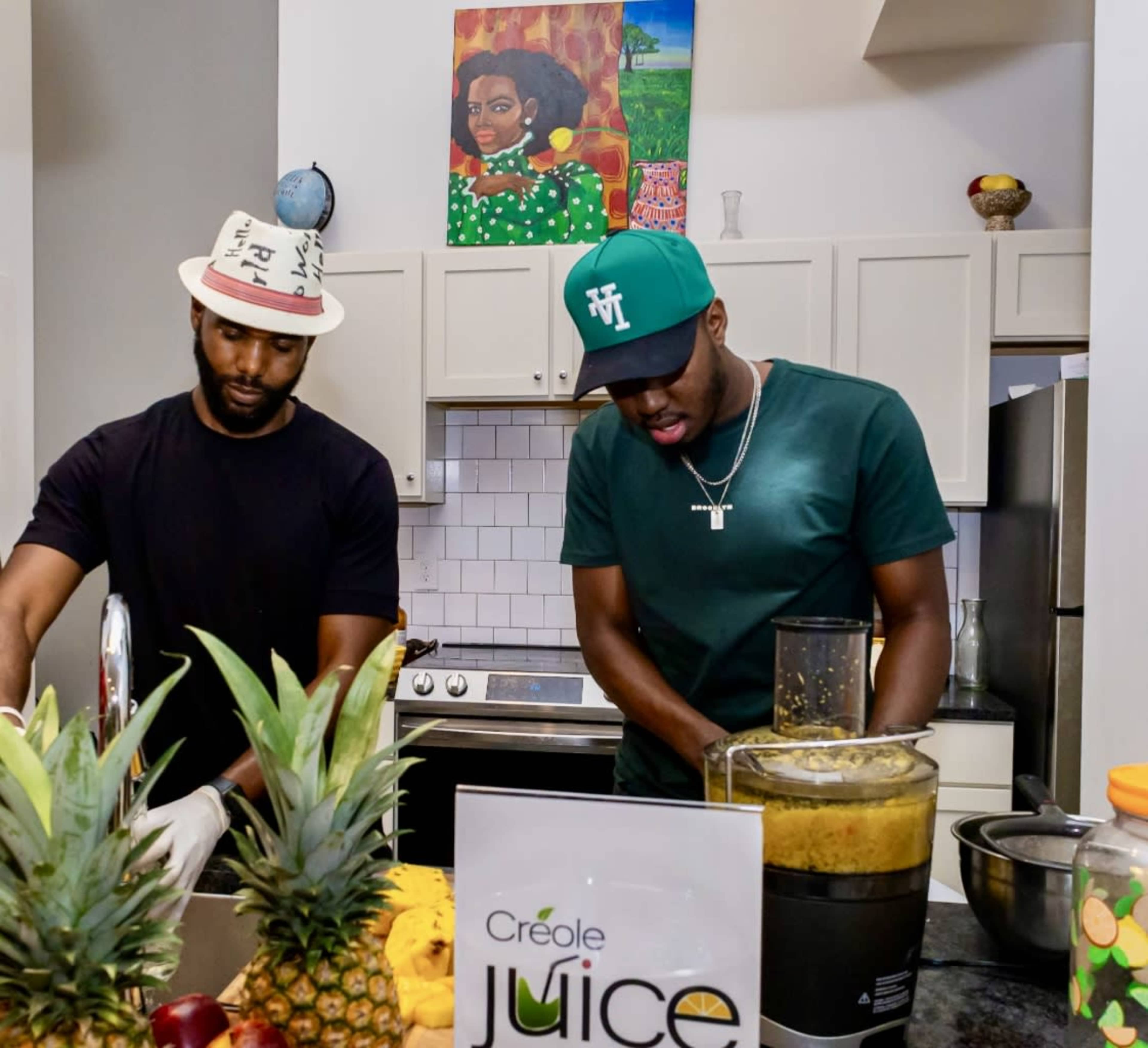 Two men are preparing juice in a kitchen, with fresh fruits and a blender in front of them.