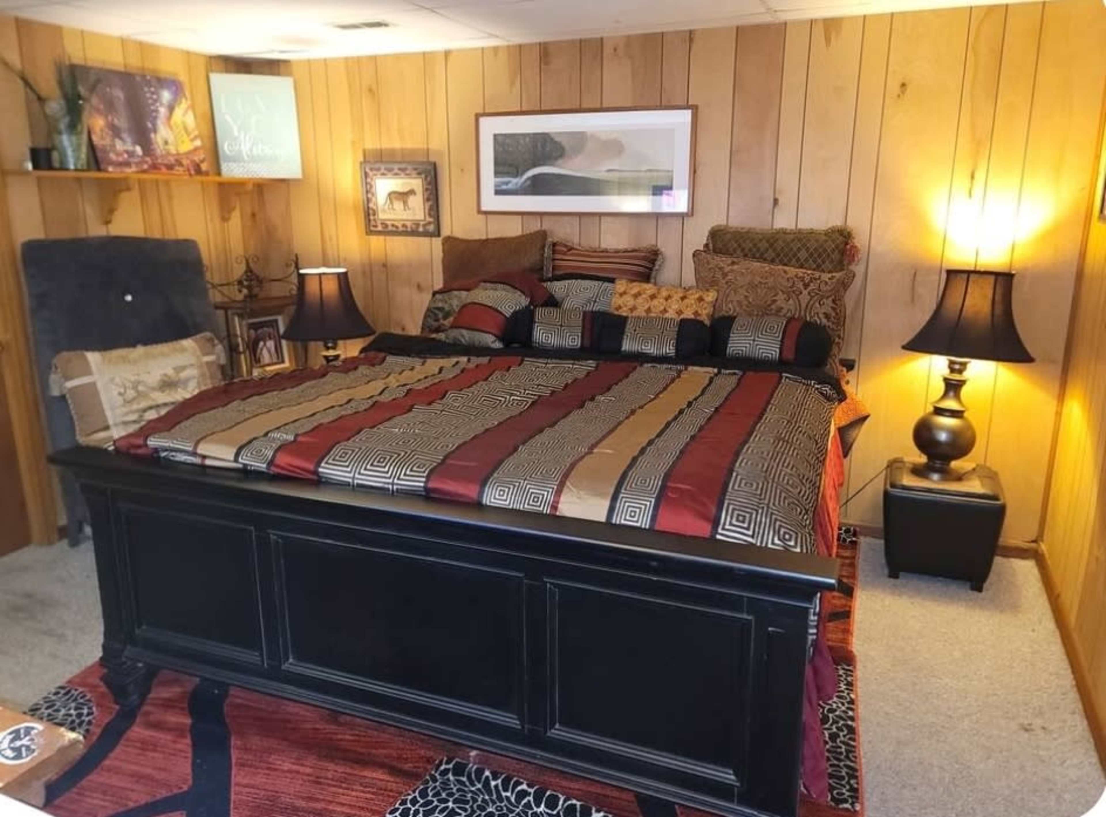 The image shows a neatly arranged bedroom featuring a large bed with decorative bedding, wooden paneling on the walls, and bedside lamps on both sides.