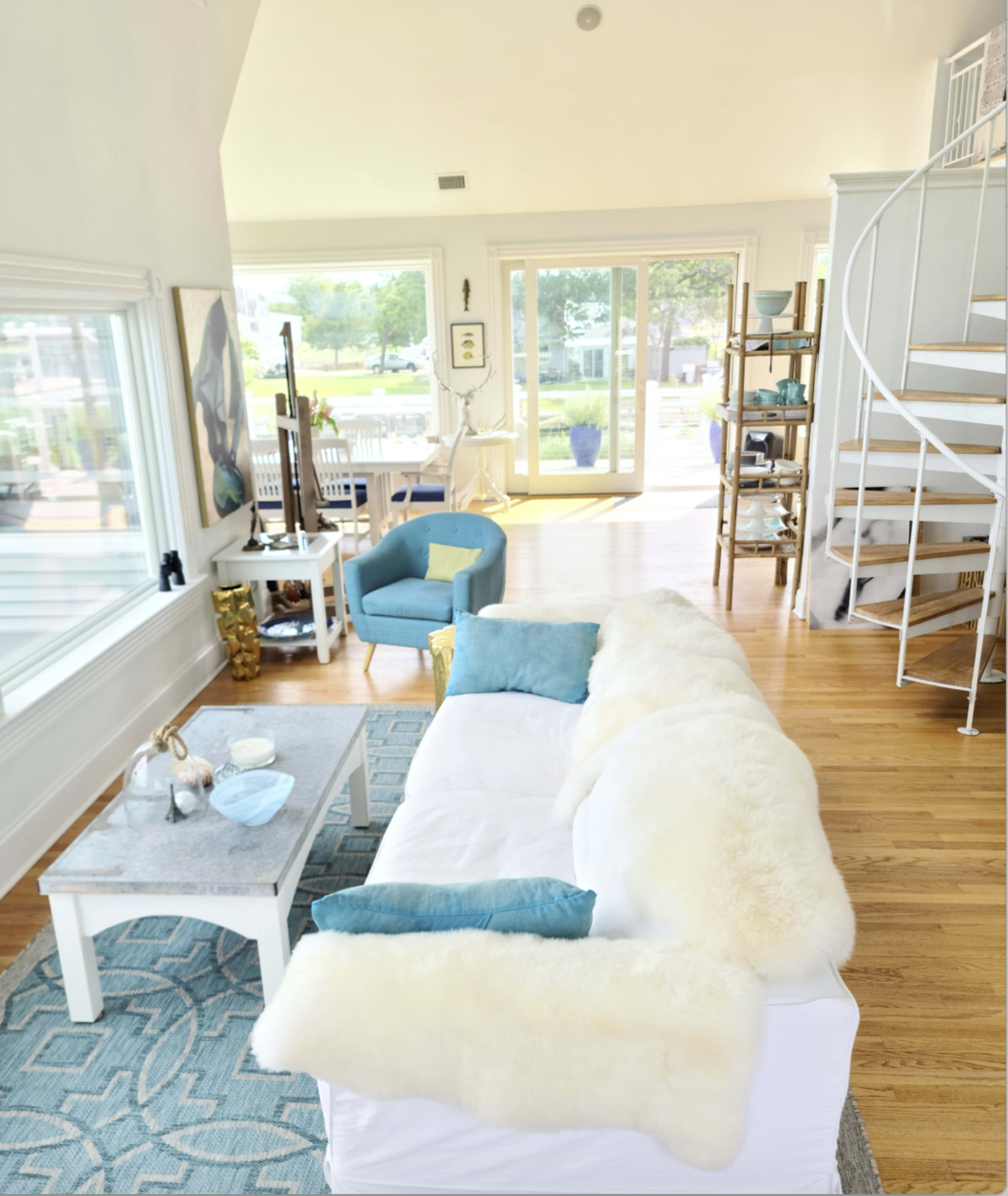 The living room features a light-colored sofa adorned with a faux fur throw, a coffee table, and a view of large windows that overlook a bright, outdoor space with spiral stairs nearby.