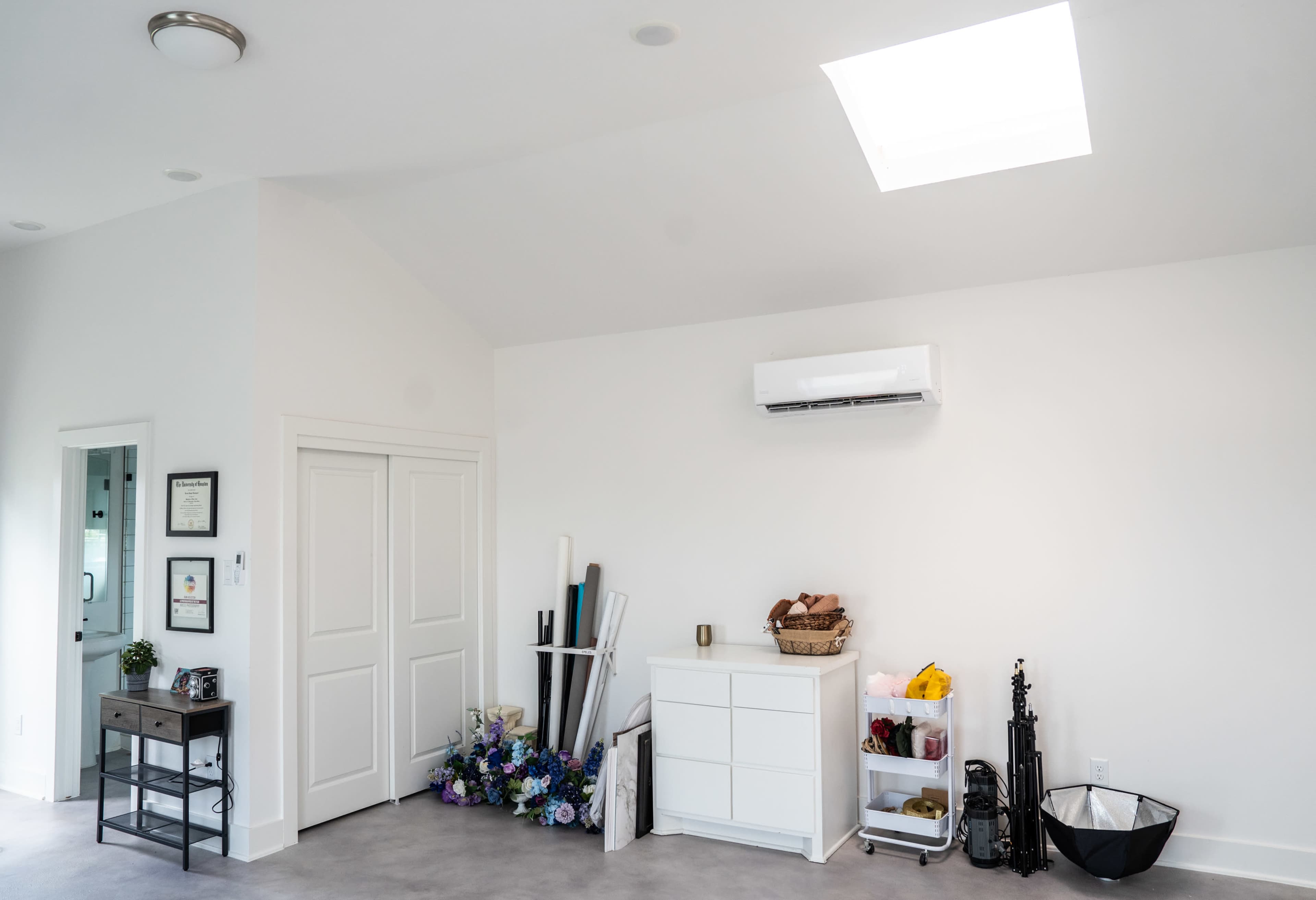 The image shows a minimalist room with a skylight, a white cabinet, a small shelf with various items, and a floral arrangement on the floor.