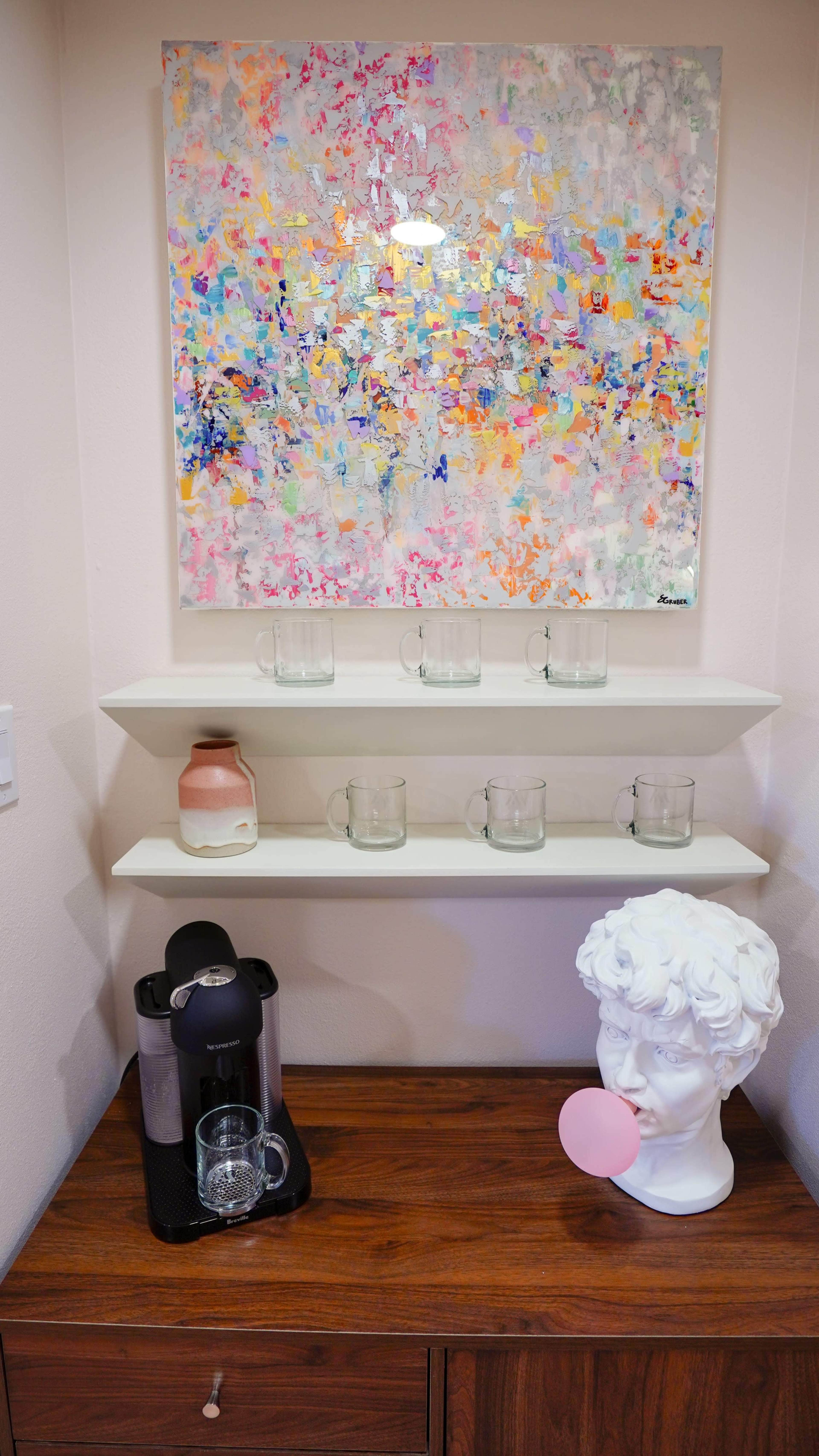 The image shows a wooden table with a coffee machine, a glass, a white sculpture of a head with a pink bubble gum, and two white shelves above displaying several glass mugs and a decorative ceramic item.