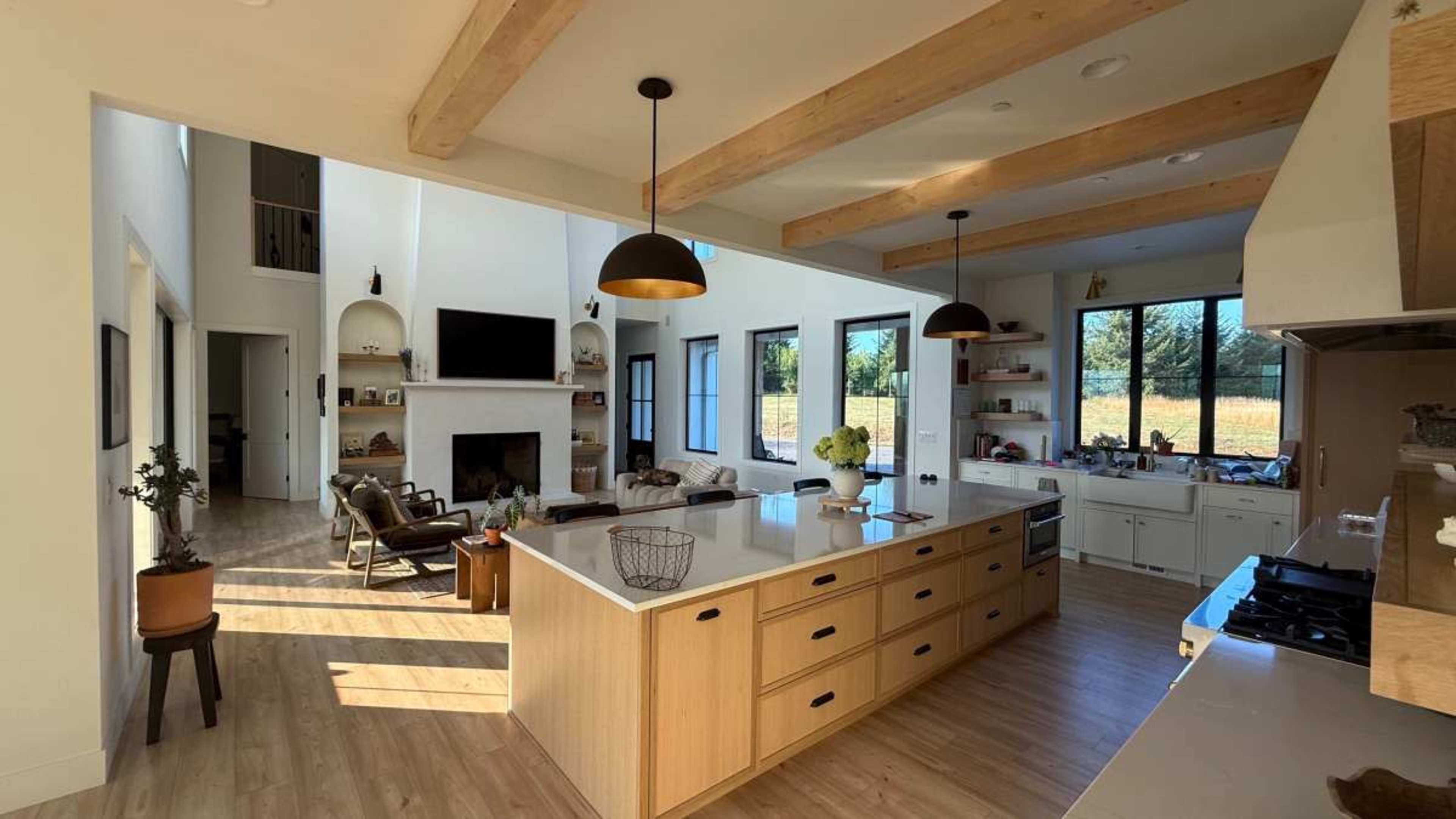 The image shows a modern kitchen with an island, wooden beams on the ceiling, and adjoining living spaces that include a fireplace and large windows.