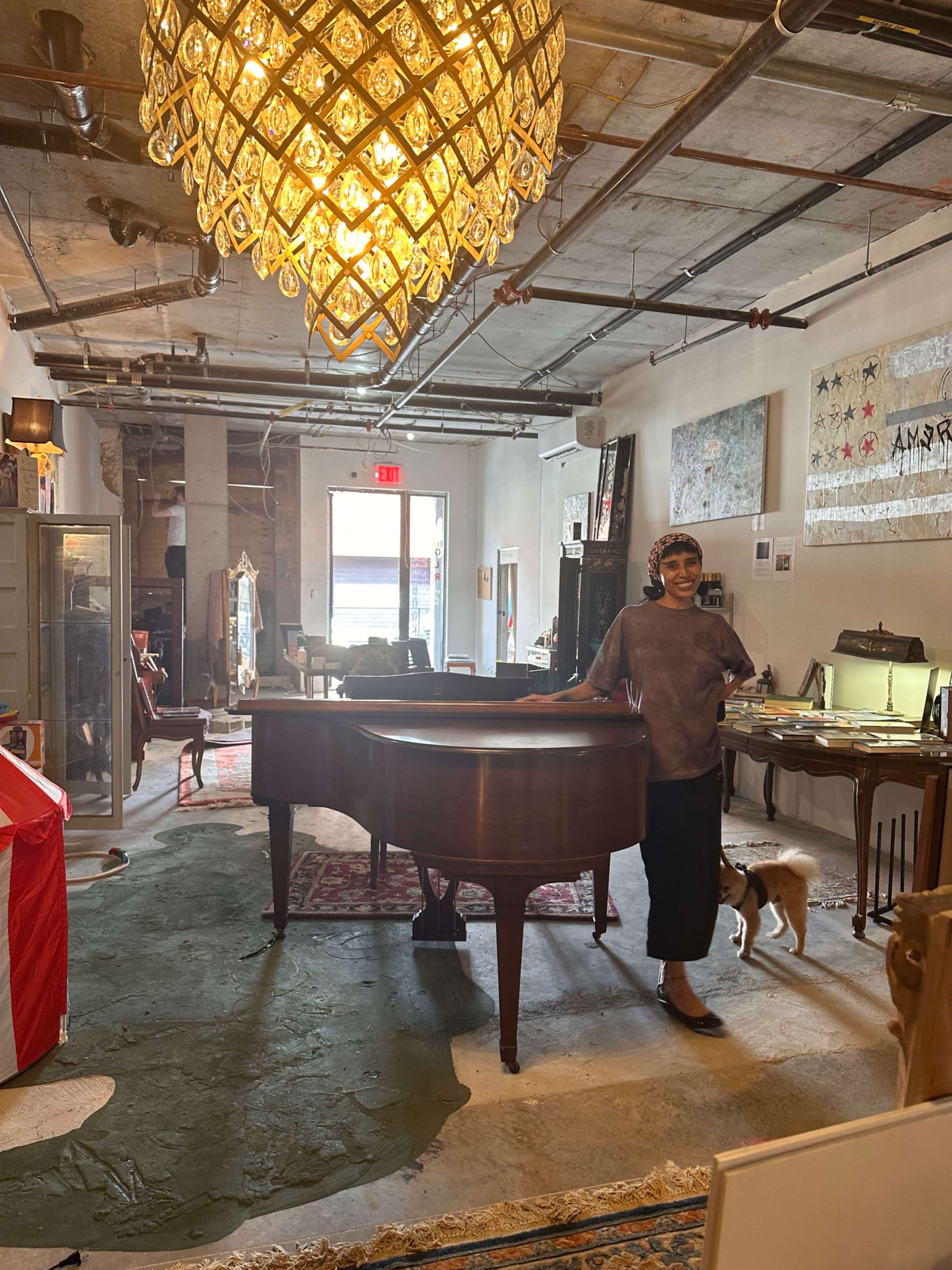 A person stands beside a grand piano in an eclectic art-filled space featuring various furnishings and a large decorative chandelier.