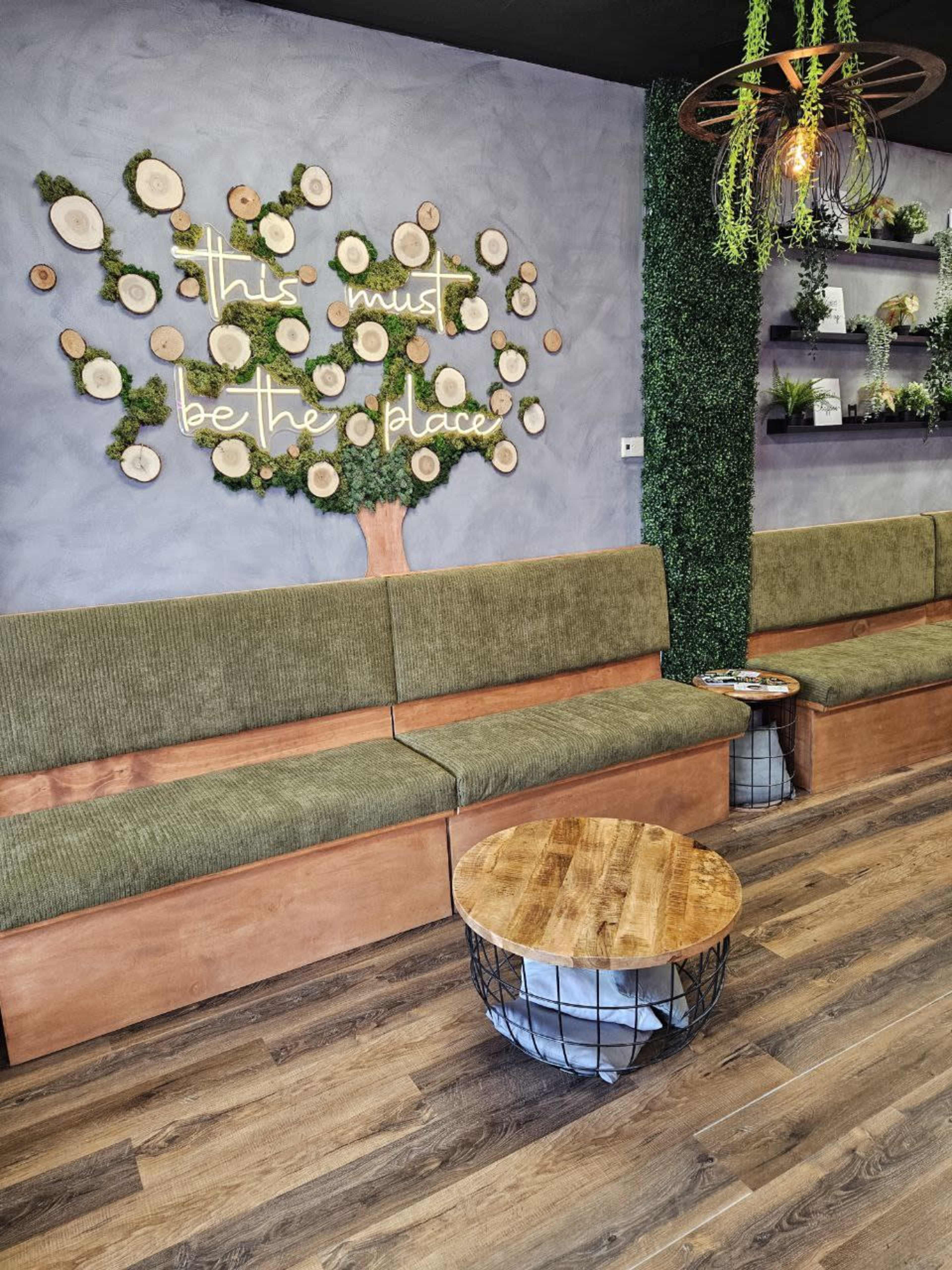 A cozy interior features green upholstered seating, a wooden table, and a decorative wall with a tree made of circular wooden slices and a neon sign.