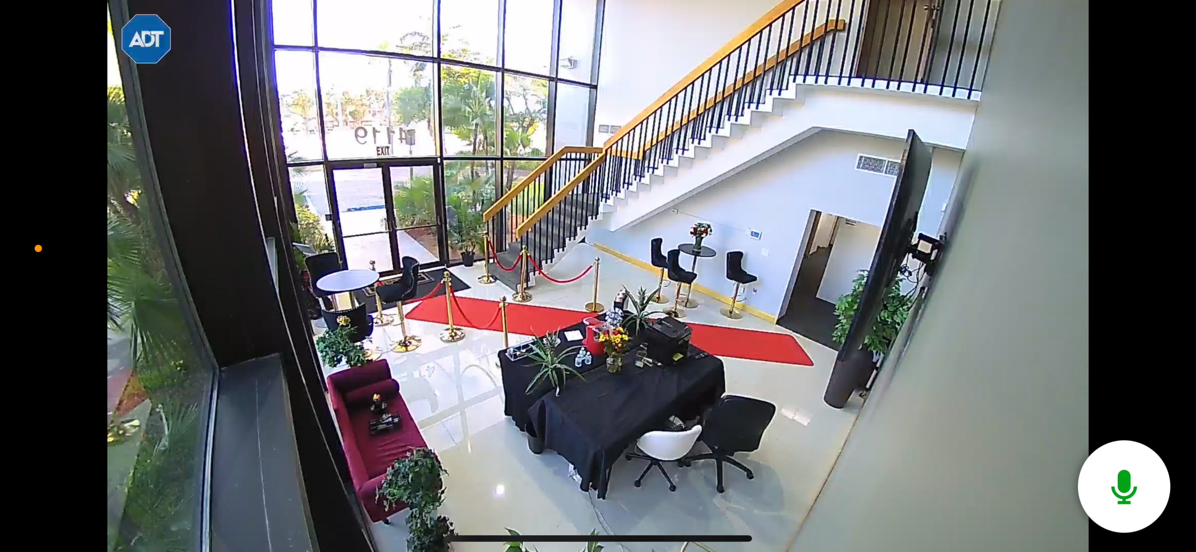 The image shows a modern lobby area with a staircase, seating arrangements, and decorative plants, featuring a red carpet leading to a reception desk.