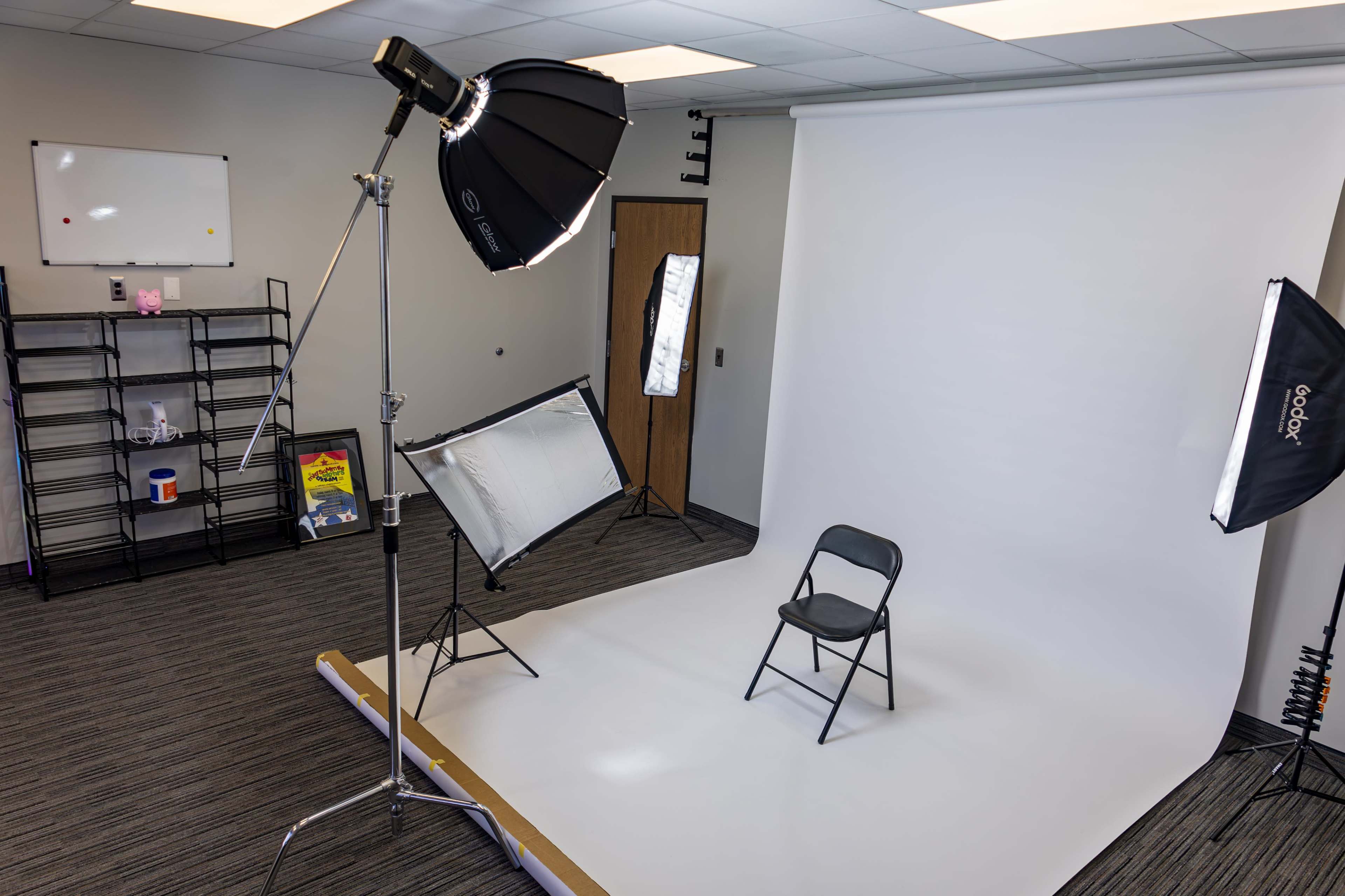 The image shows a photography studio setup featuring a chair on a white backdrop, with lighting equipment positioned around the area.