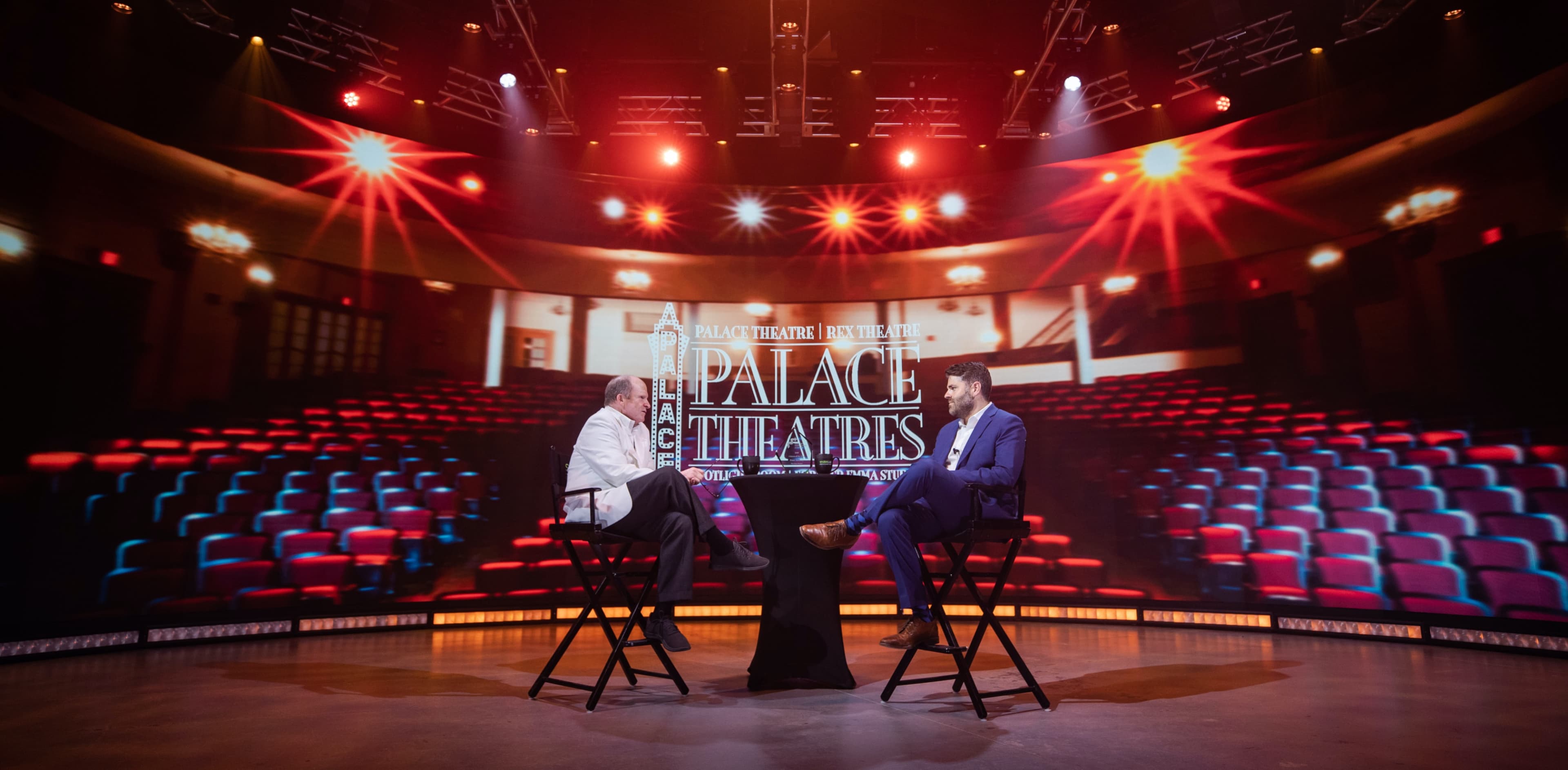 A stage is set for an interview, featuring two seated individuals in front of a large screen displaying "Palace Theatres" with images of an empty theater behind them.