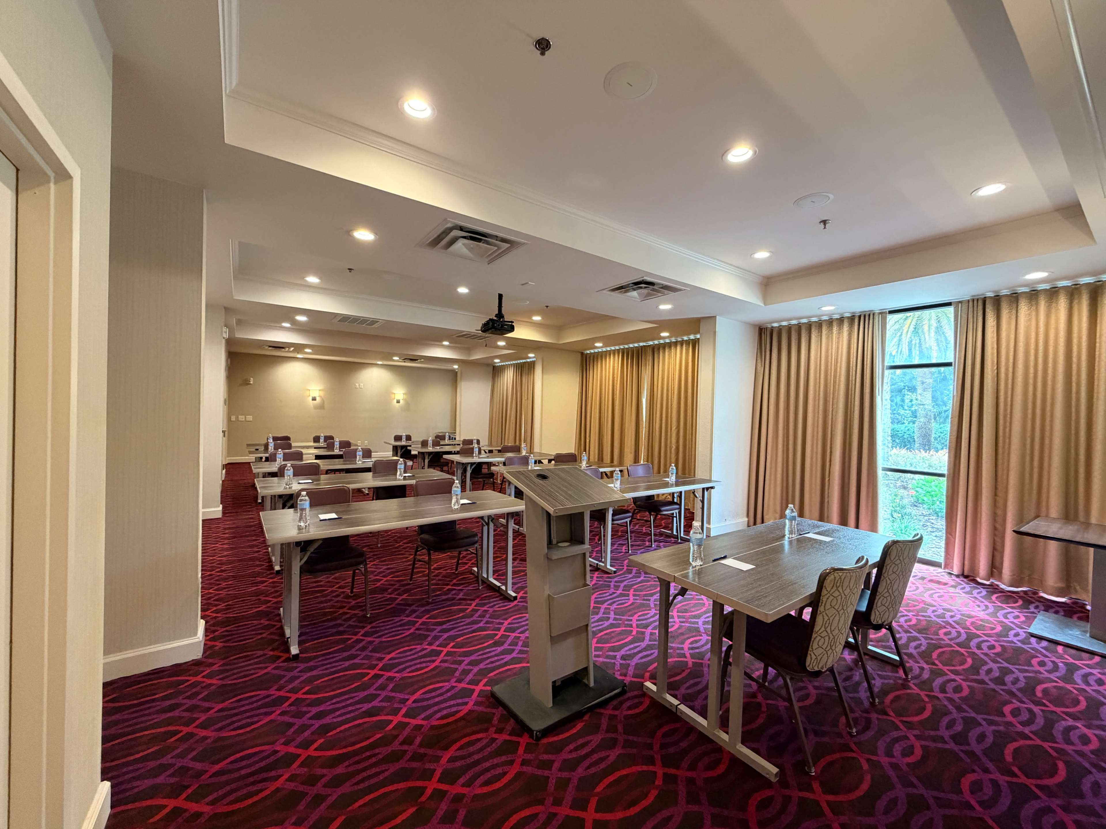 The image shows a well-lit conference room arranged with several tables and chairs, featuring a podium in the foreground and large windows with curtains in the background.