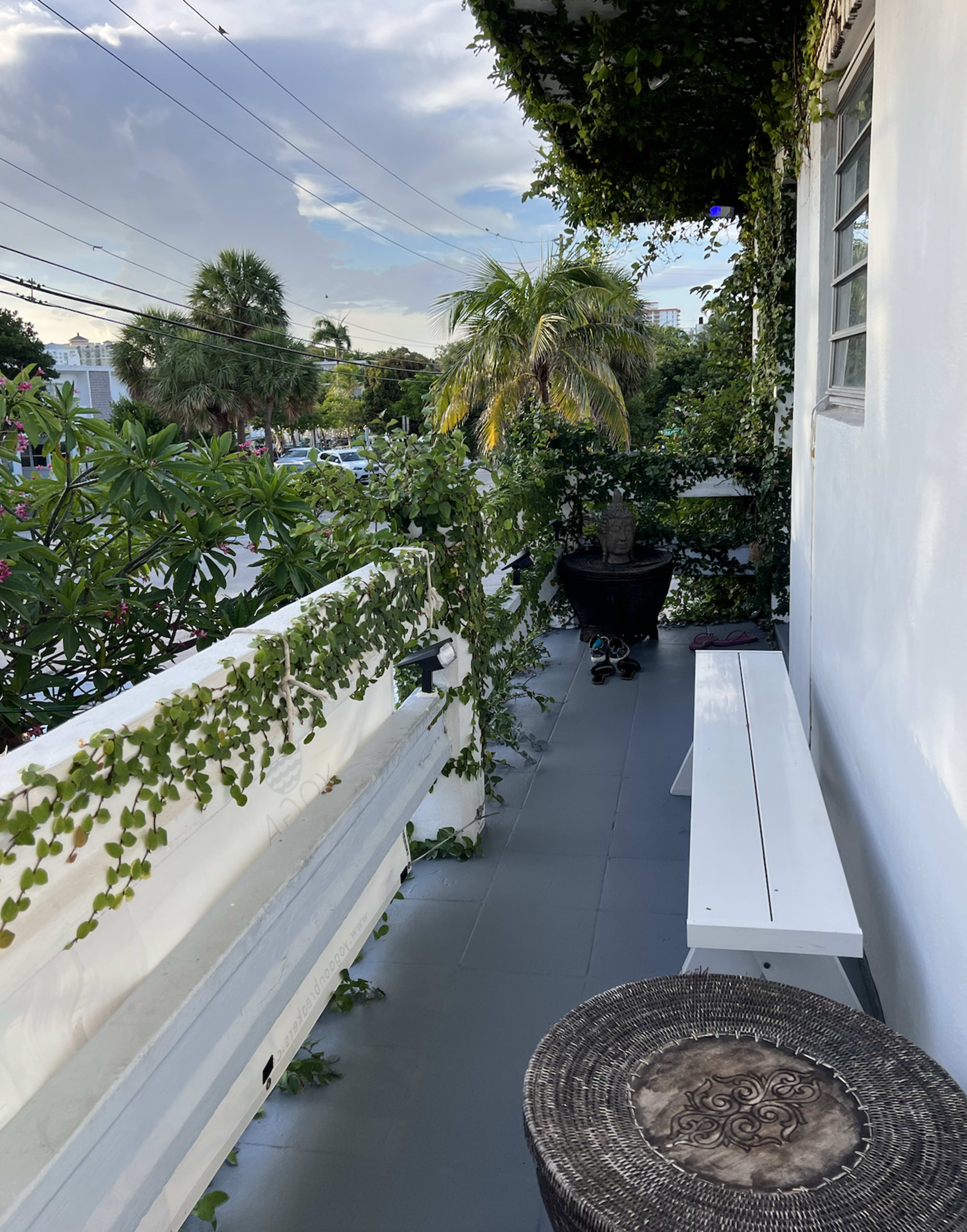 The image shows a narrow balcony adorned with green plants, featuring a white bench and a round stool, with palm trees visible in the background.
