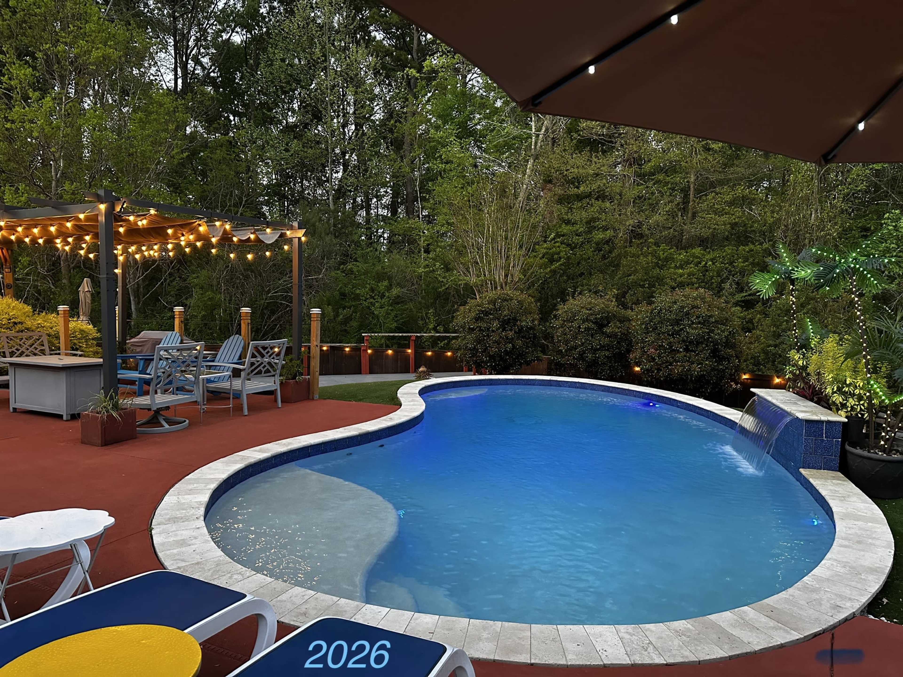 The image shows a backyard pool area with a curved pool, a wooden pergola with seating, and surrounding greenery illuminated by string lights.