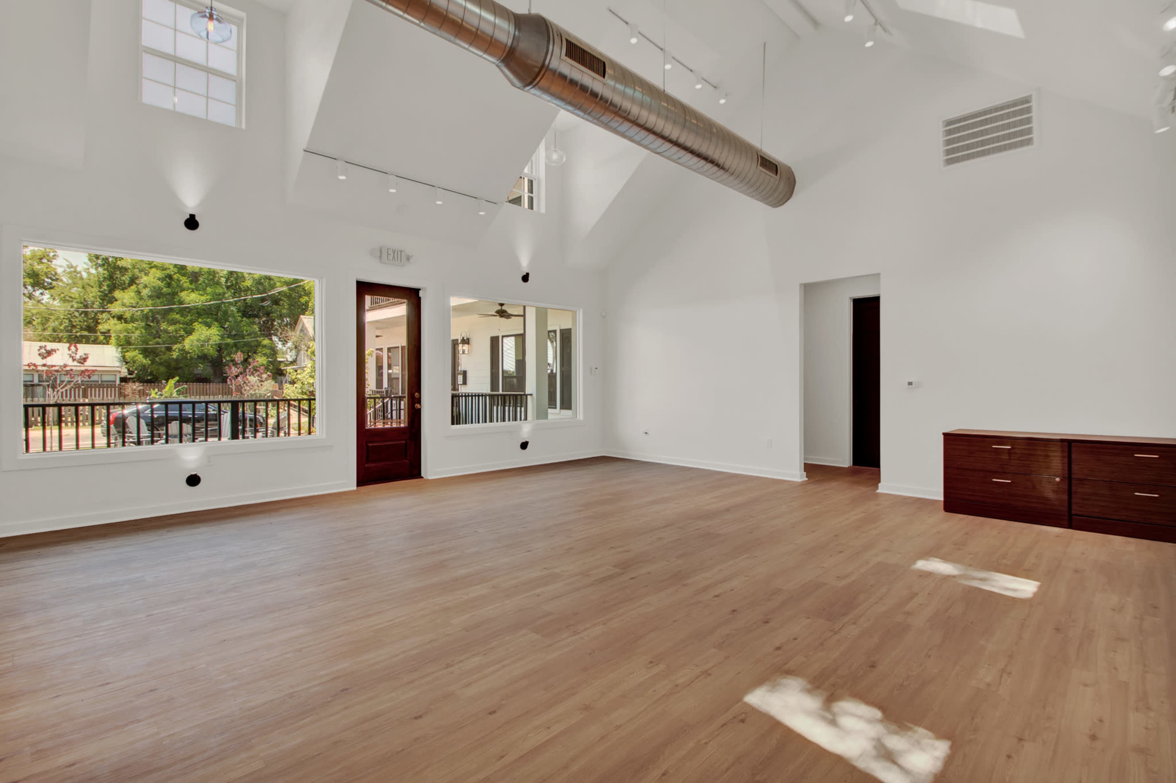 The image shows a bright, spacious room with wooden flooring, large windows, and minimal furniture.