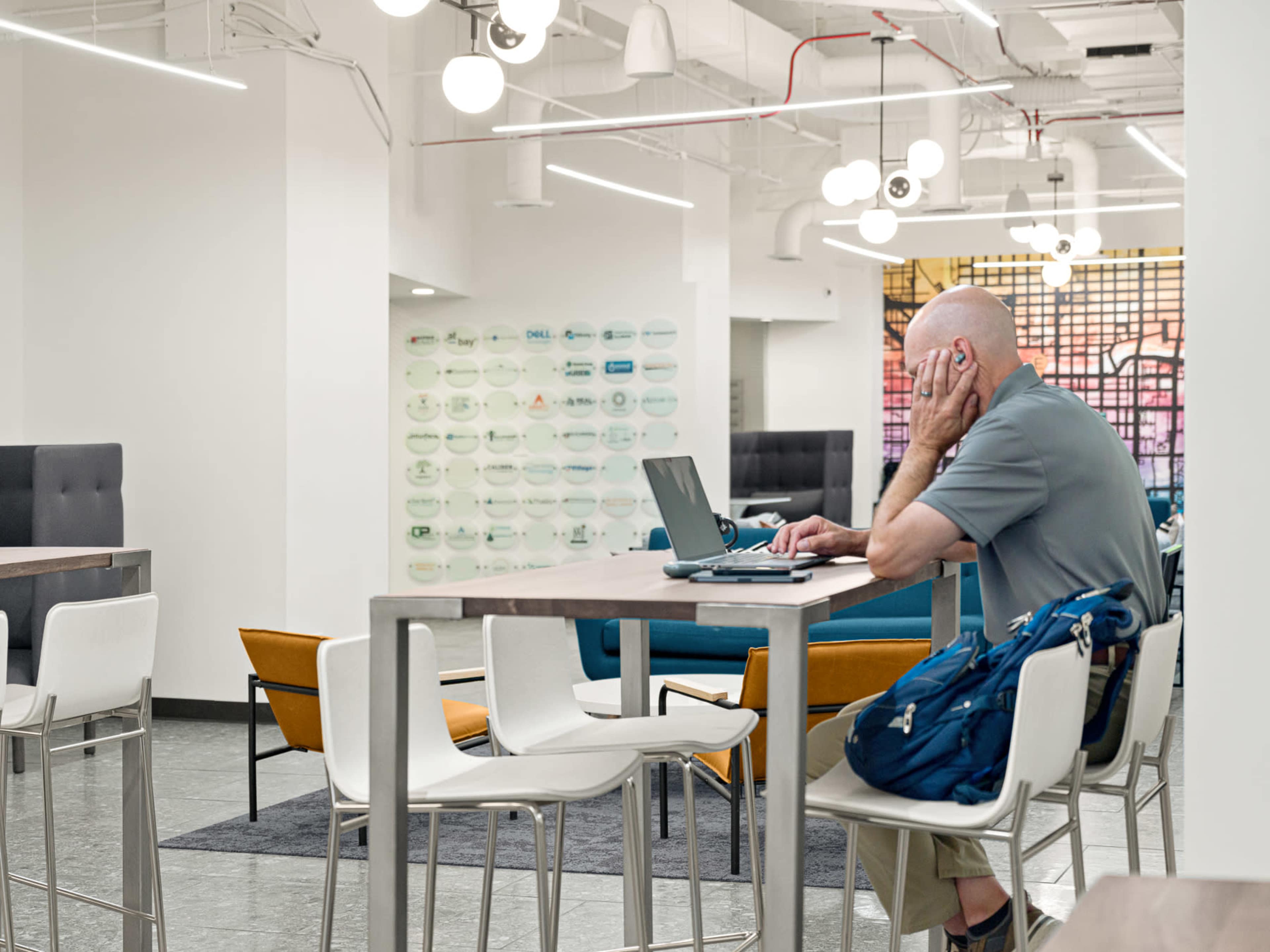 A bald man sits at a high table working on a laptop in a modern, open office space with multiple seating areas.