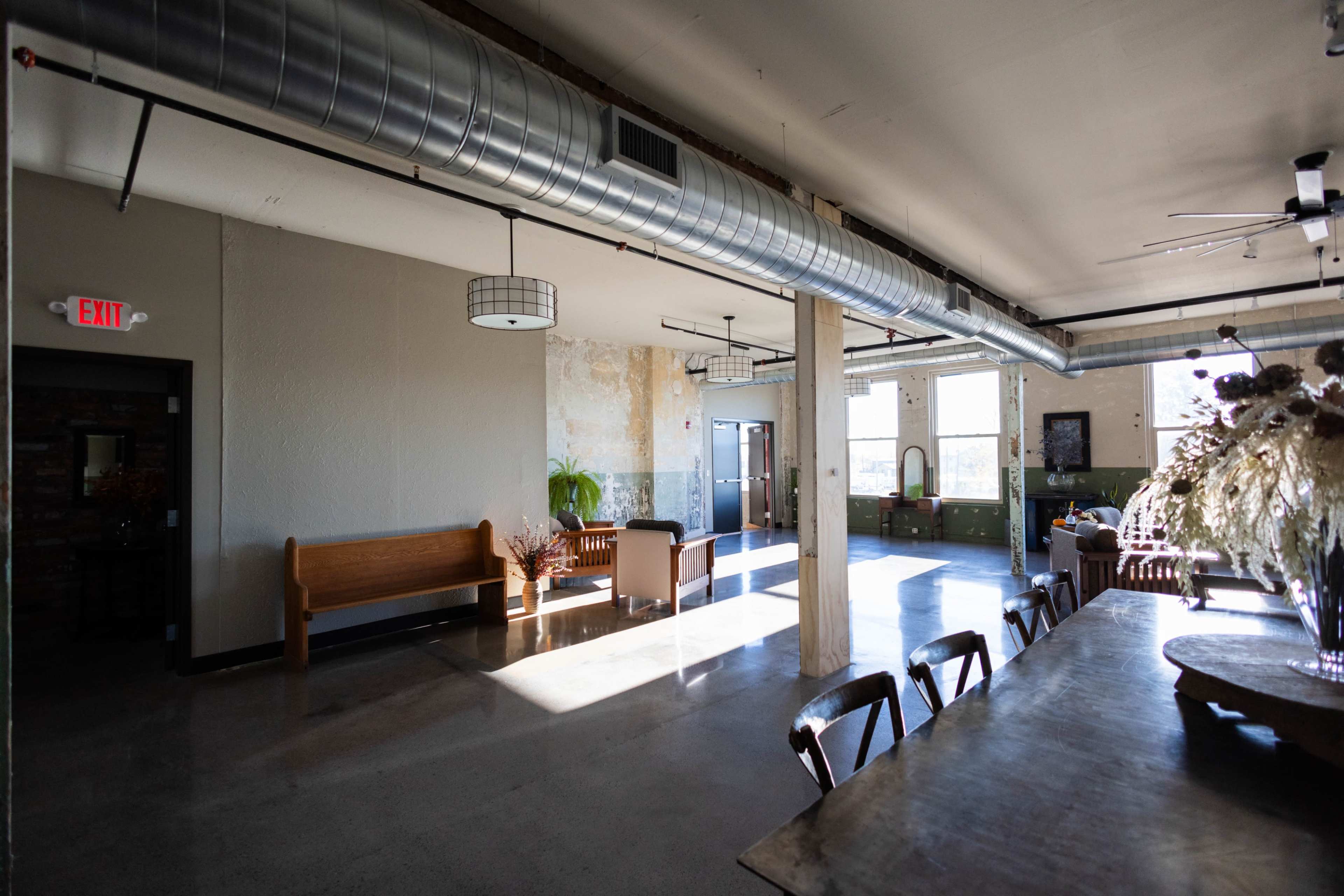 A spacious room with industrial-style decor, featuring a long dining table, benches, and large windows letting in natural light.