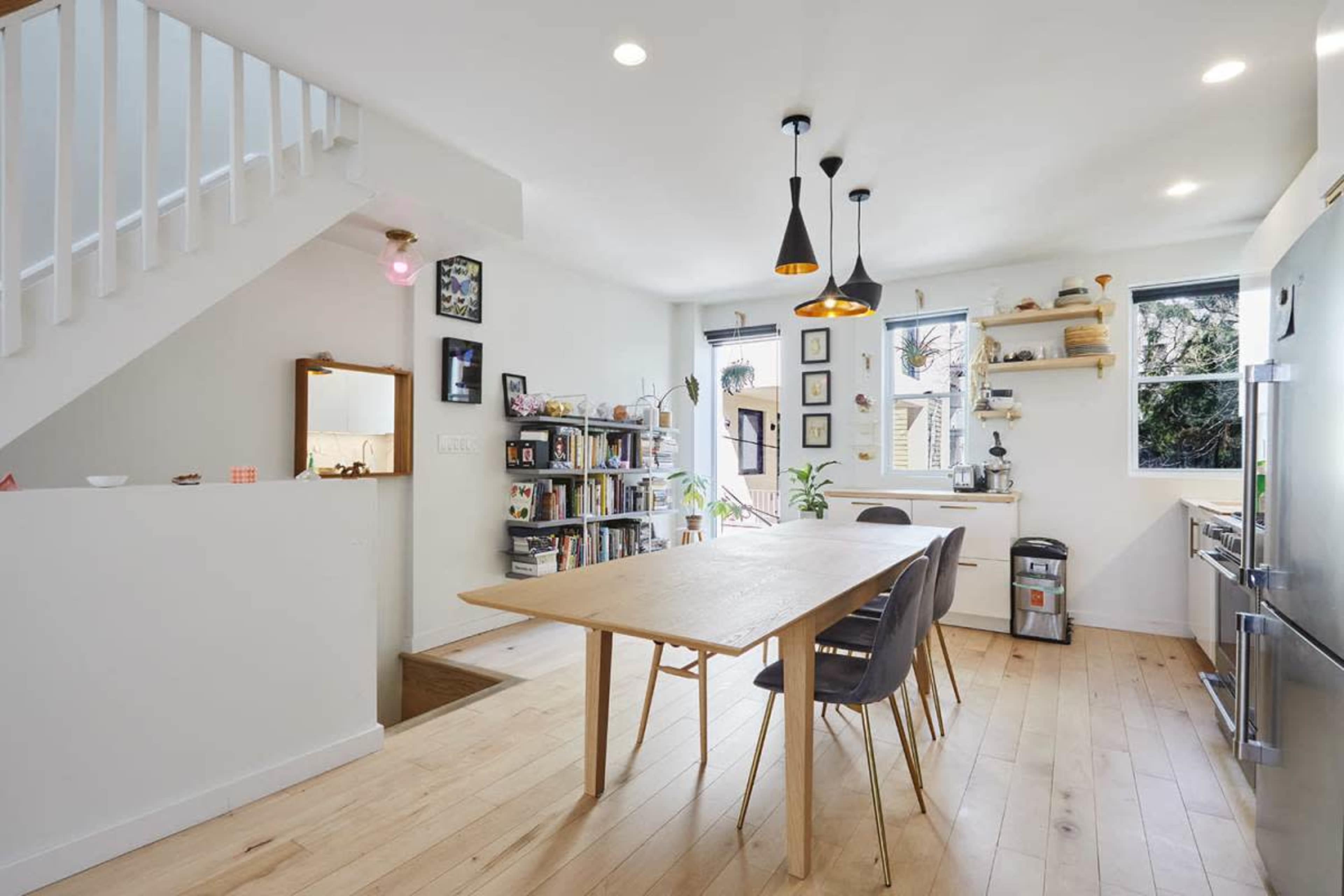 A bright, modern kitchen features a large wooden table surrounded by black chairs, with shelves of books and decorative items lining the walls.