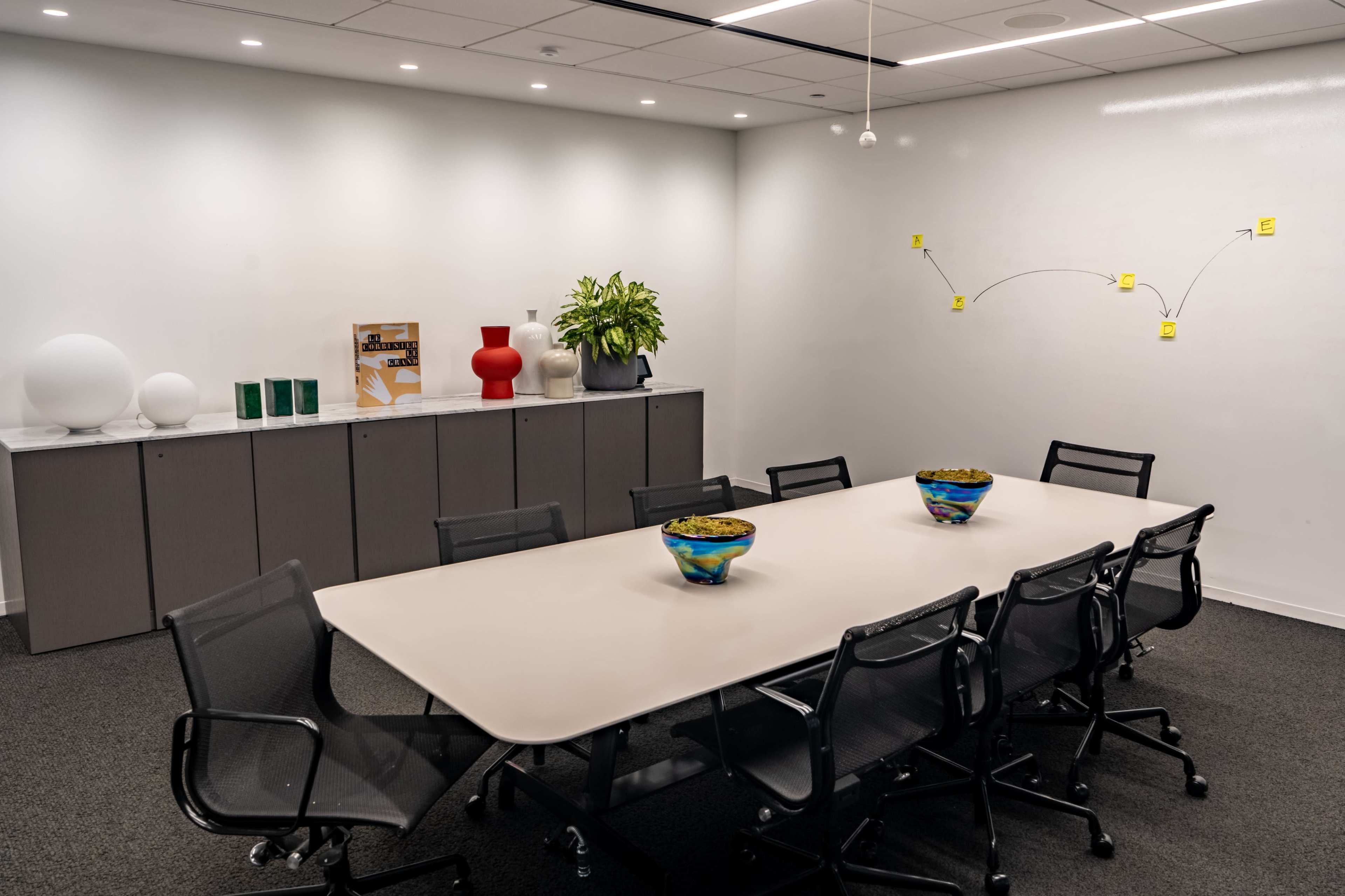 A modern conference room features a large table surrounded by black chairs, with a side cabinet displaying decorative items and a whiteboard on the wall.