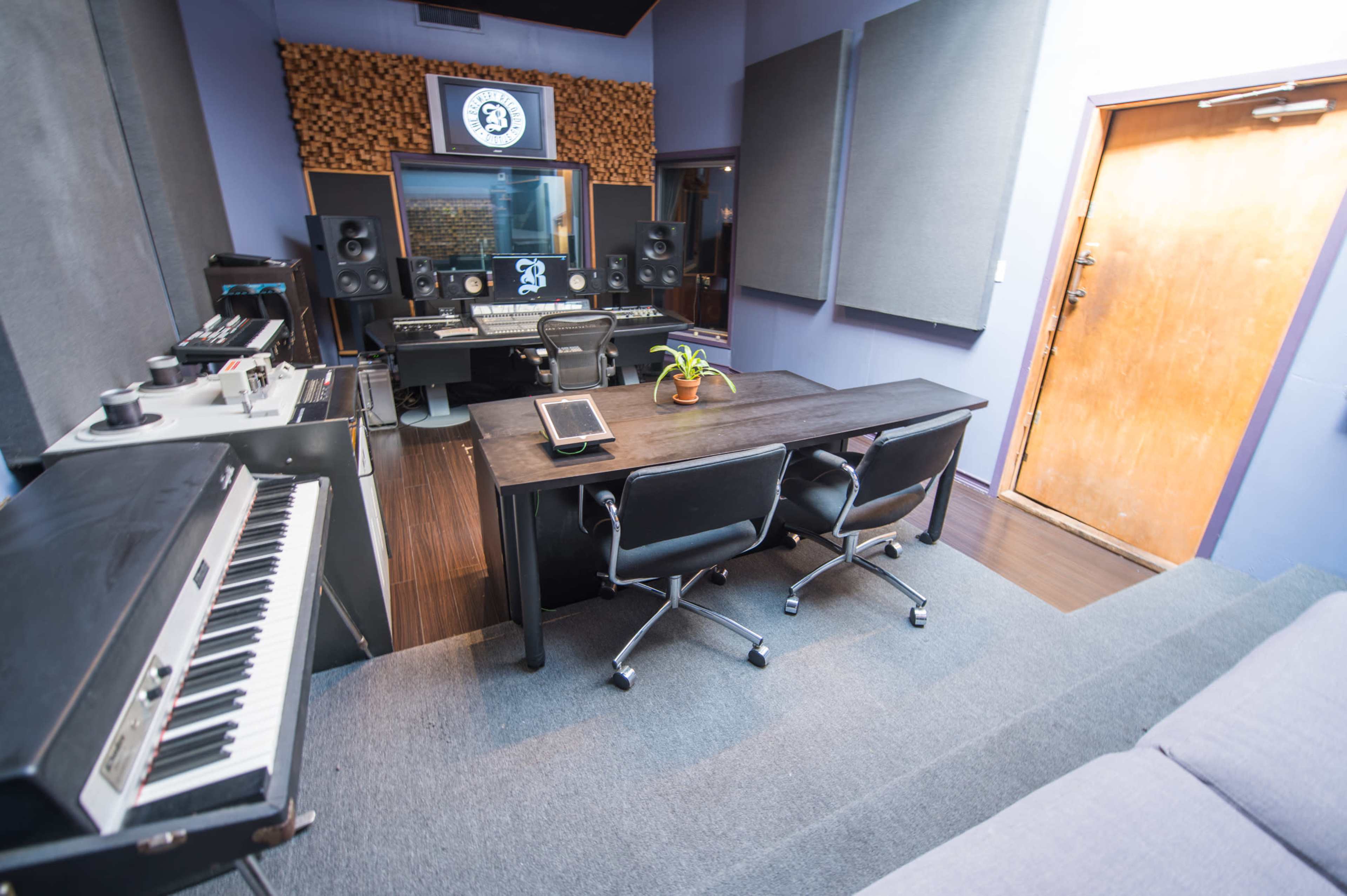 A music studio with a keyboard, a large mixing desk, two chairs, soundproofing panels on the walls, and a wooden door.