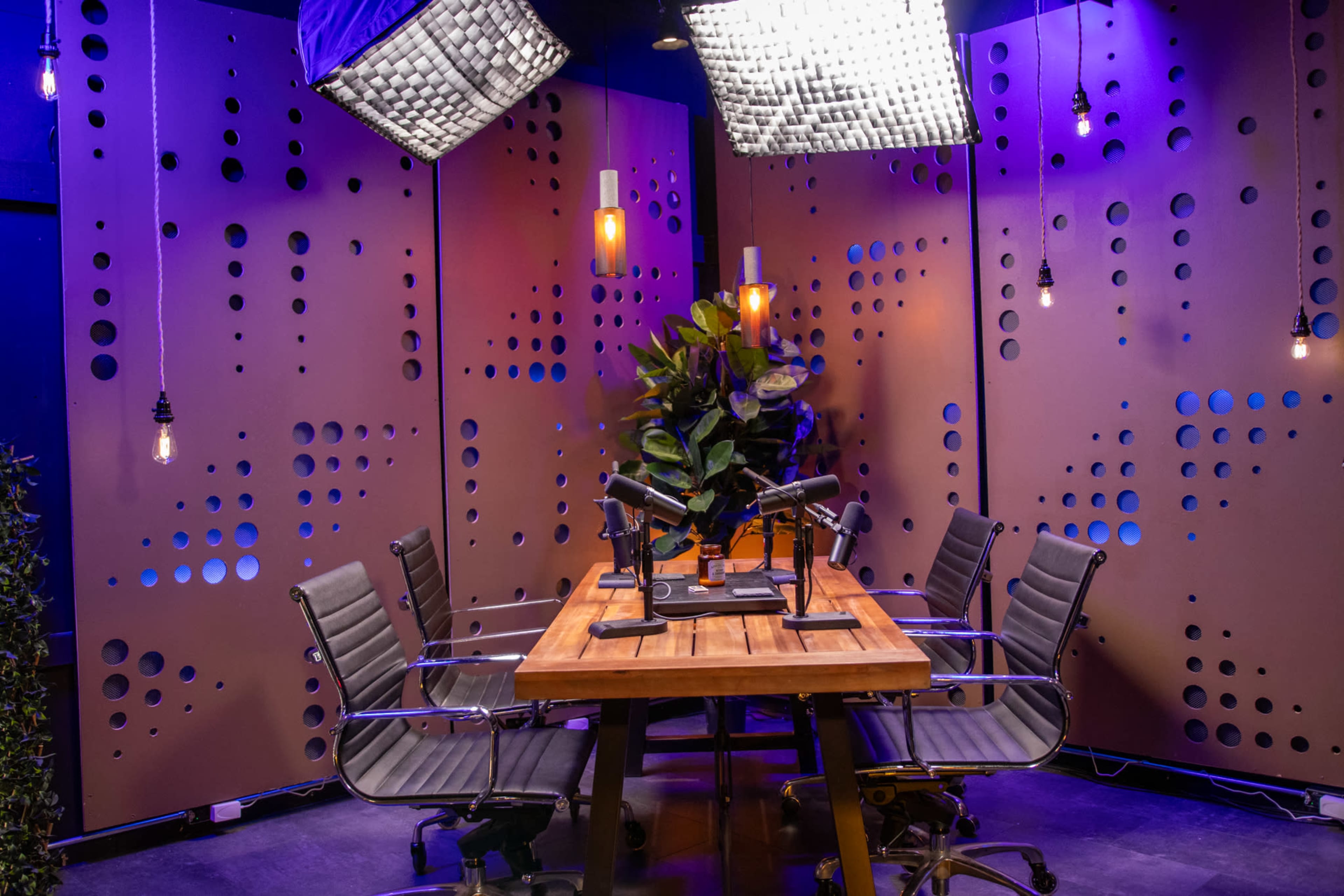 A modern podcast studio features a wooden table surrounded by sleek chairs, with microphones set up and overhead lights illuminating the space.