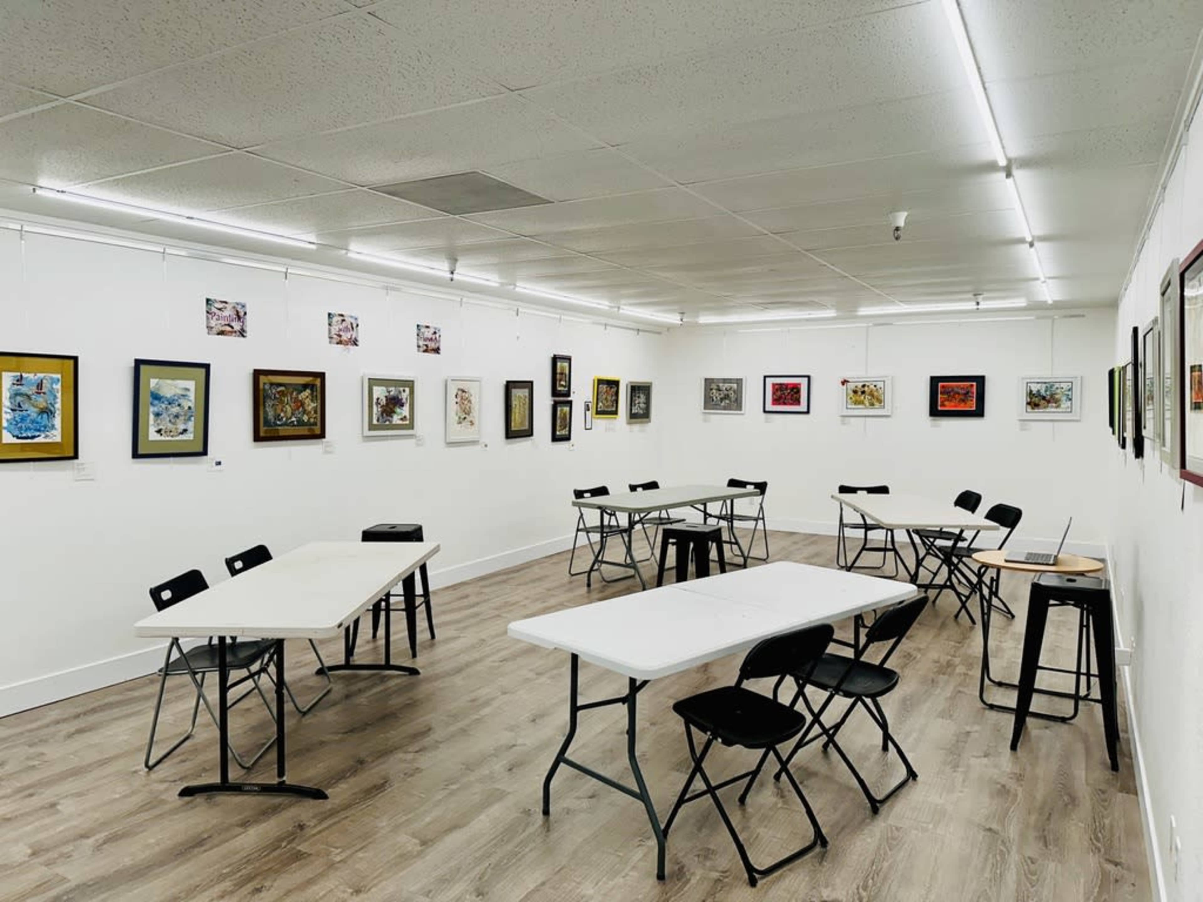 An art gallery with several framed artworks displayed on the walls and multiple tables and chairs arranged in the center.