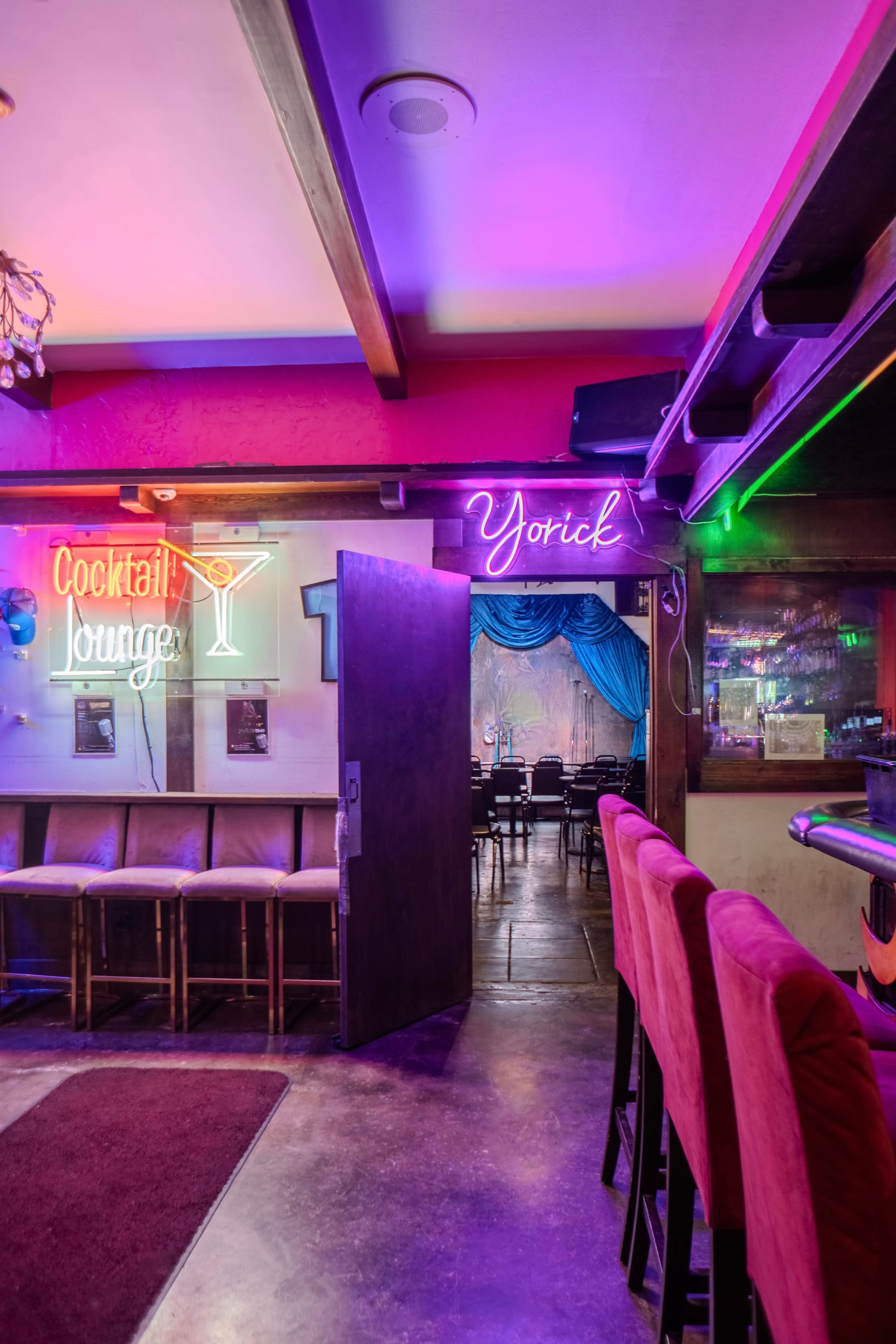 A bar area with neon signs for "Cocktail Lounge" and "Yorick," leading into a dimly lit room adorned with blue drapes.