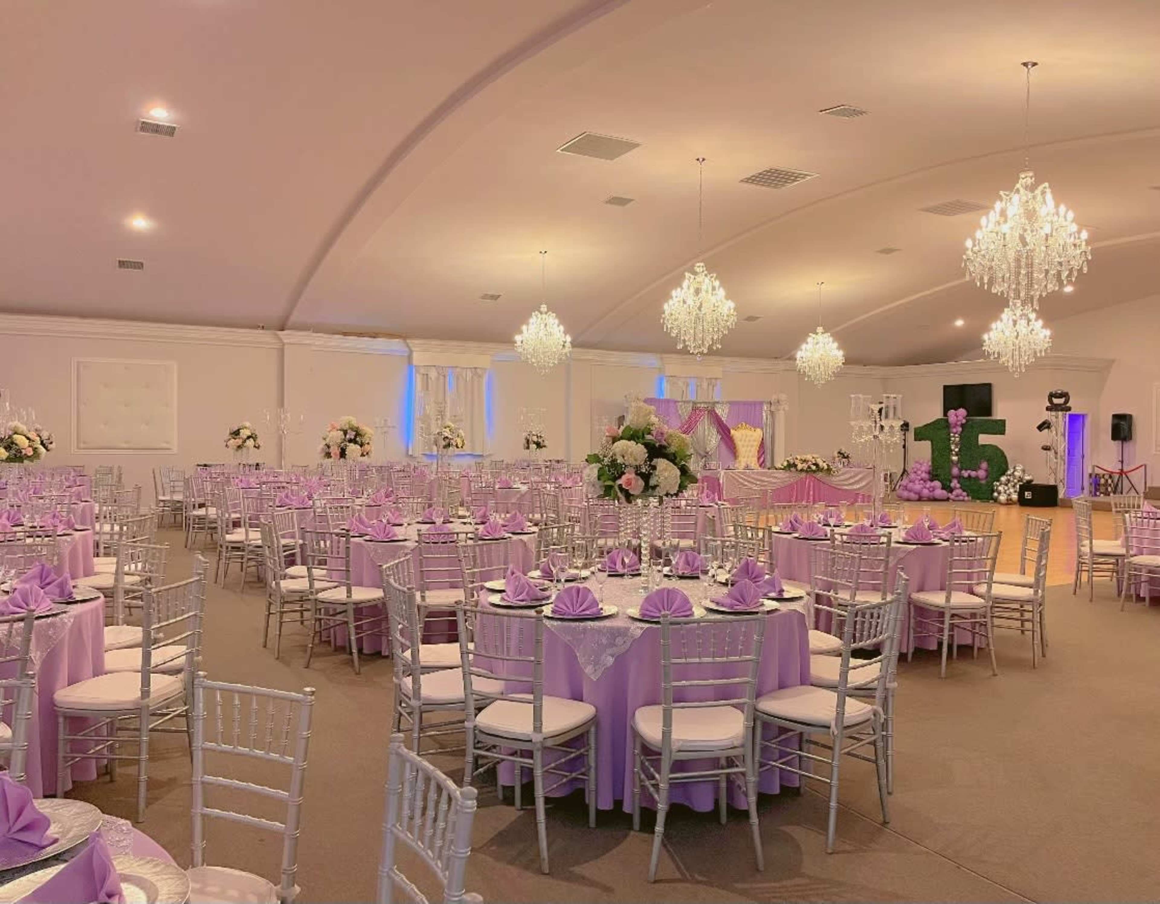 The image shows a banquet hall decorated for a special event, with round tables covered in purple linens and set with white chairs, while chandeliers illuminate the space.