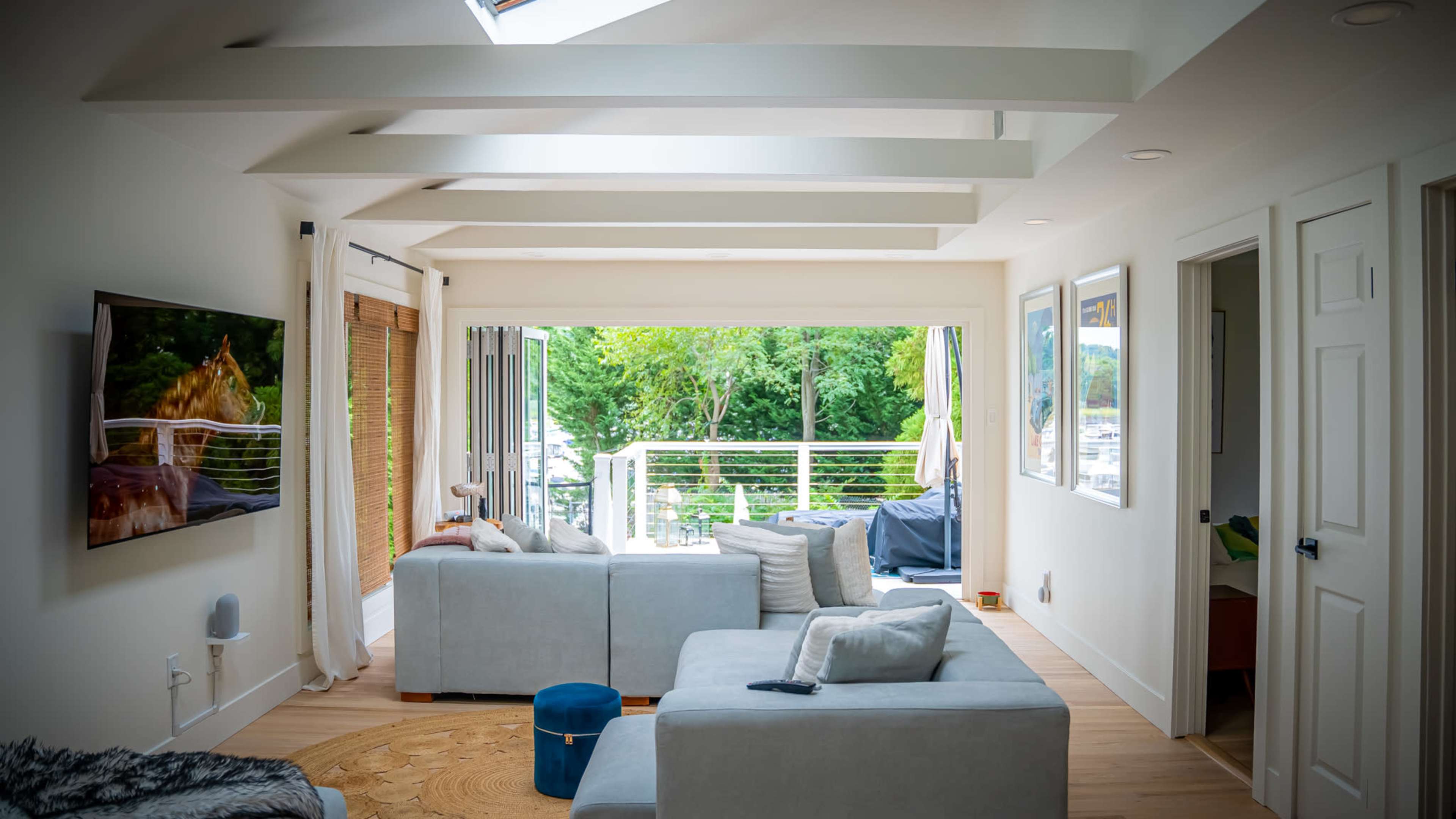 A modern living room features a light-colored sofa, a television, and large windows leading to a view of greenery outside.