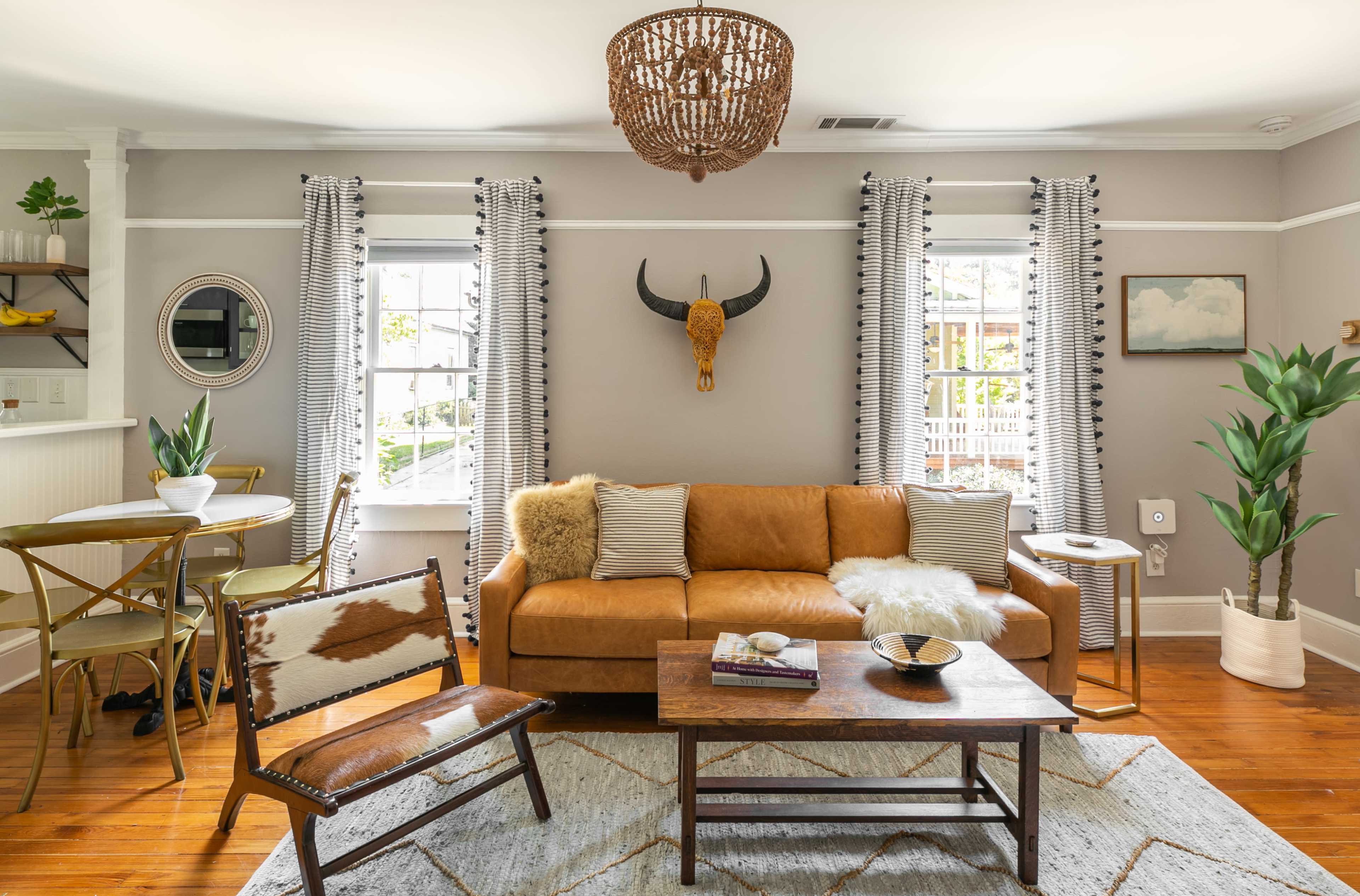 The living room features a brown leather sofa, a wooden coffee table, and a vintage chair, with large windows dressed in striped curtains and a decorative bull skull mounted on the wall.