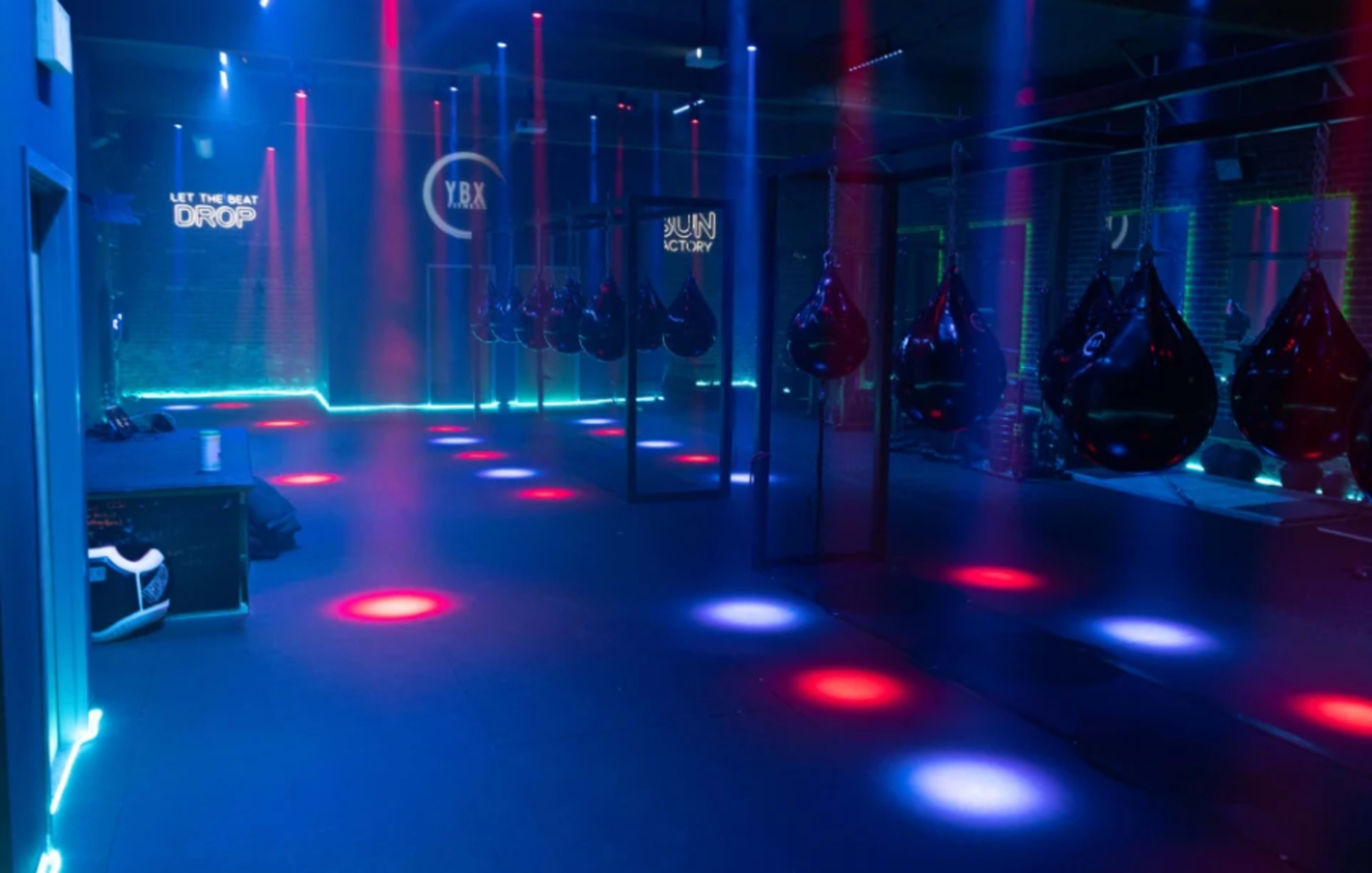 The image shows an illuminated fitness studio with hanging punching bags, colored lights, and a stage area.