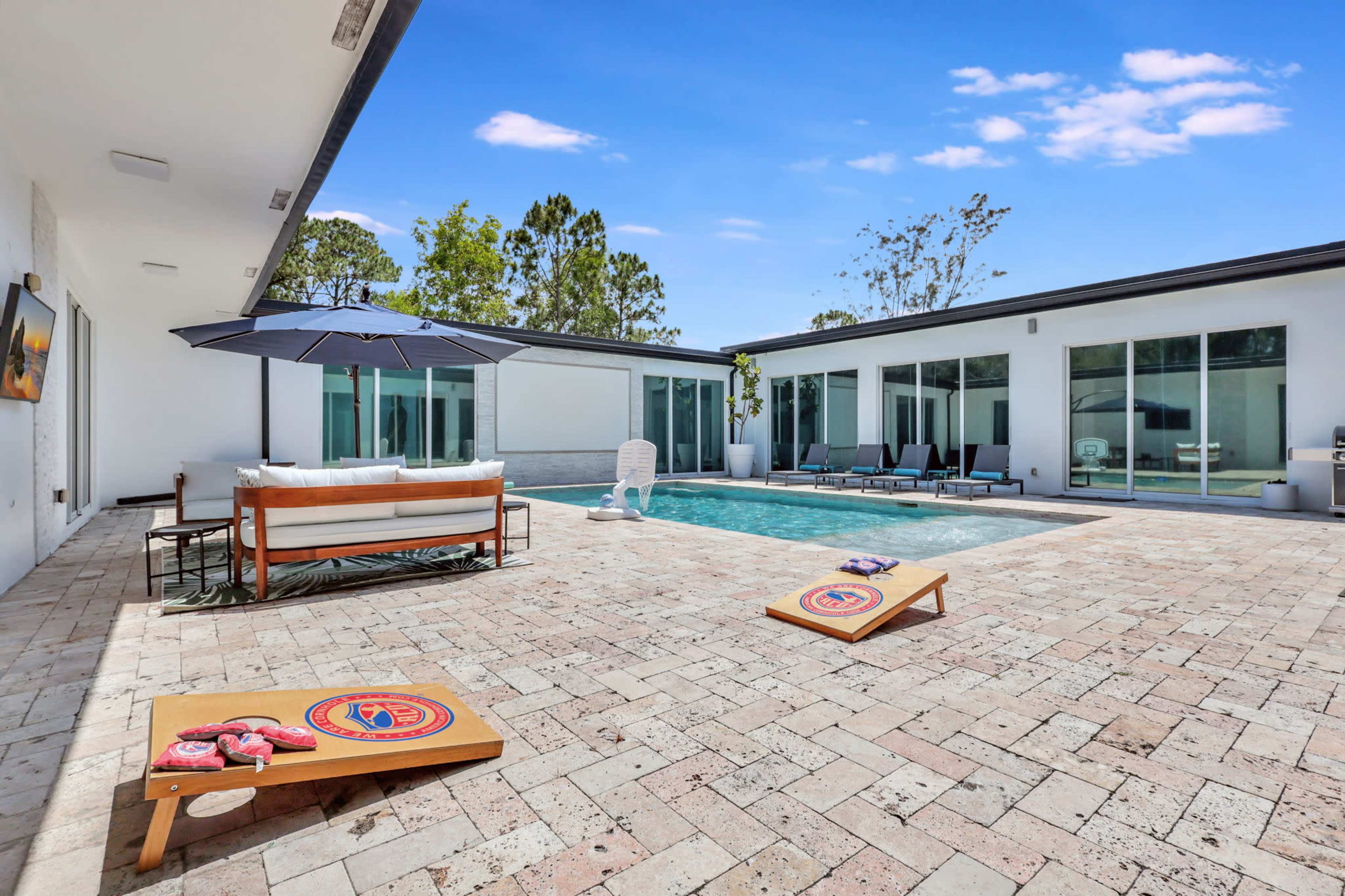 A modern outdoor space features a swimming pool surrounded by lounge chairs, a shaded seating area, and cornhole boards on a stone patio.