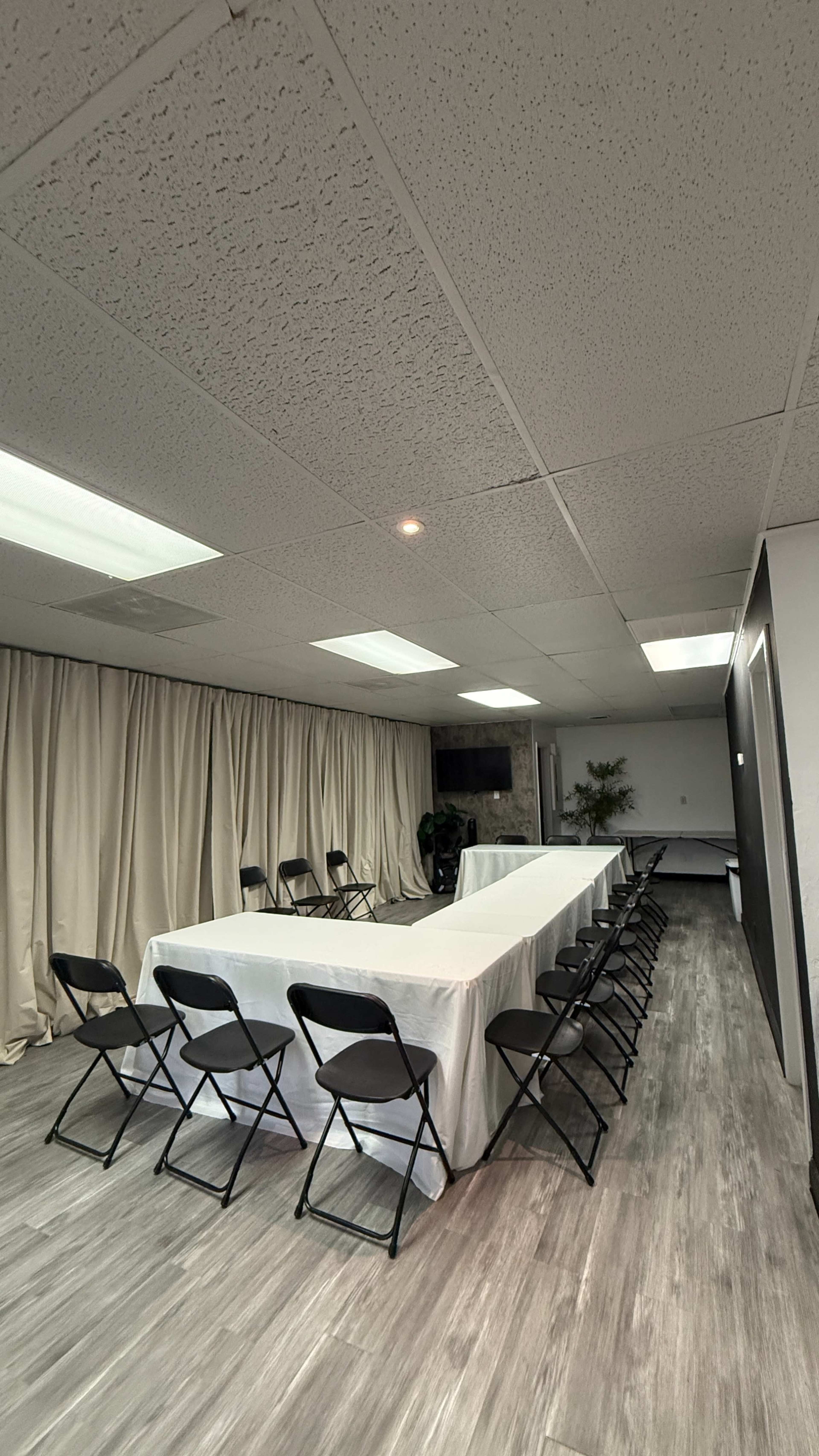 The image shows a meeting room with a long table covered by a white cloth, surrounded by black folding chairs, and beige curtains along one side.