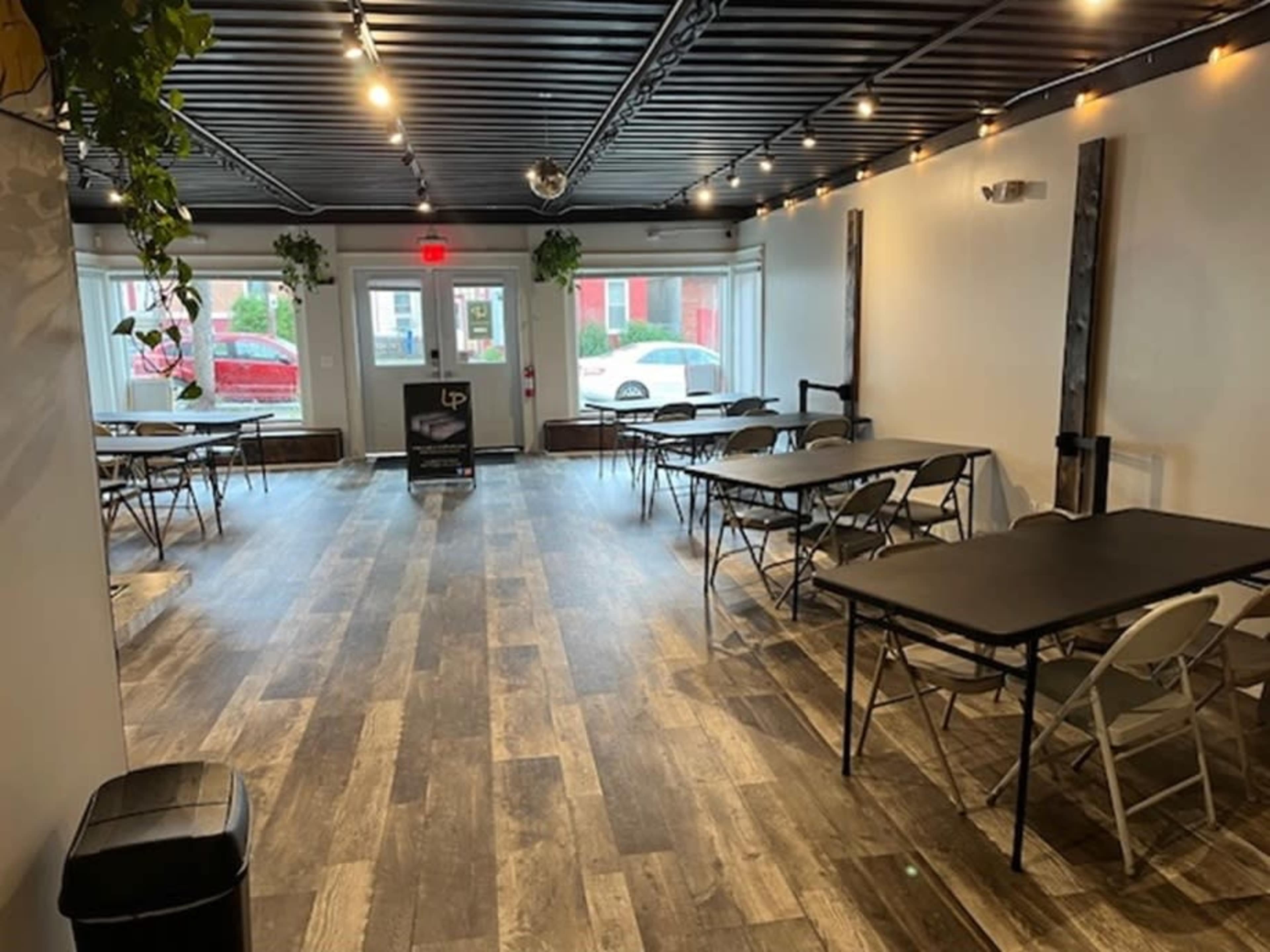 A spacious room with several rows of folding tables and chairs, illuminated by ceiling lights and featuring wooden flooring.