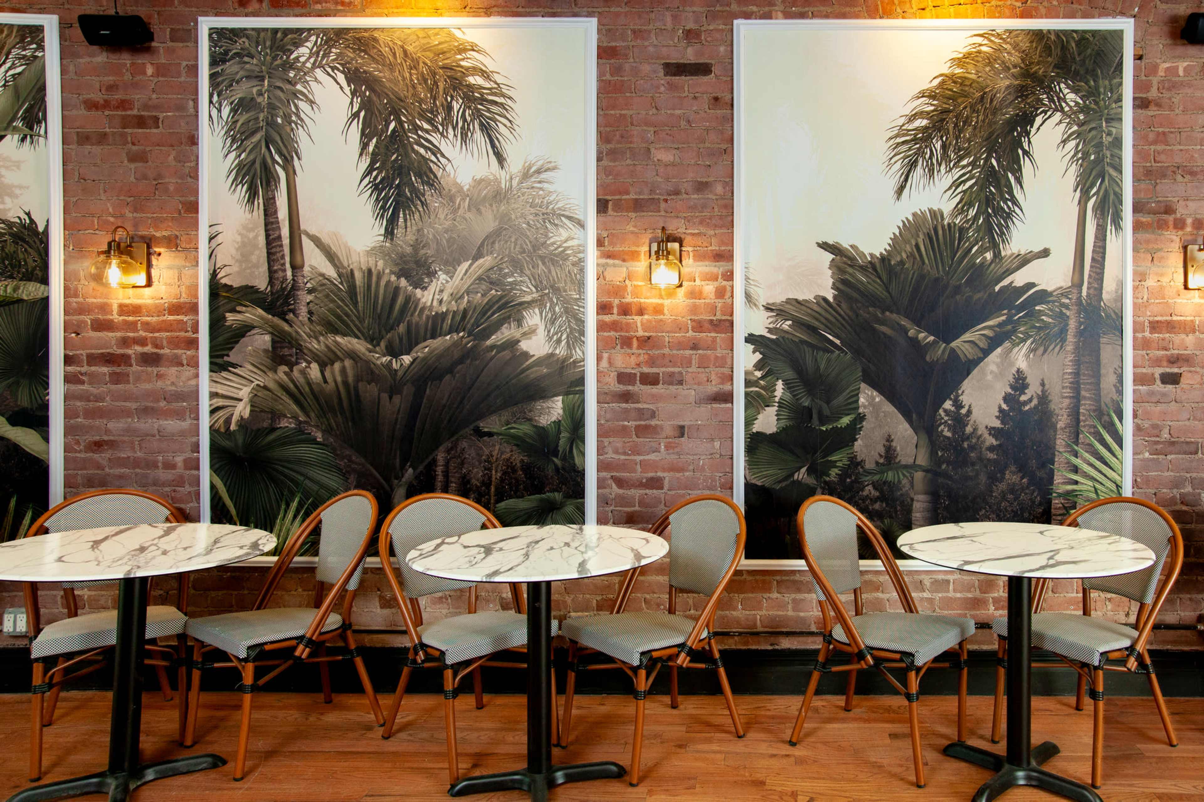 A restaurant interior featuring two large botanical-themed prints on exposed brick walls, with several marble-top tables and bamboo-style chairs arranged in front of them.