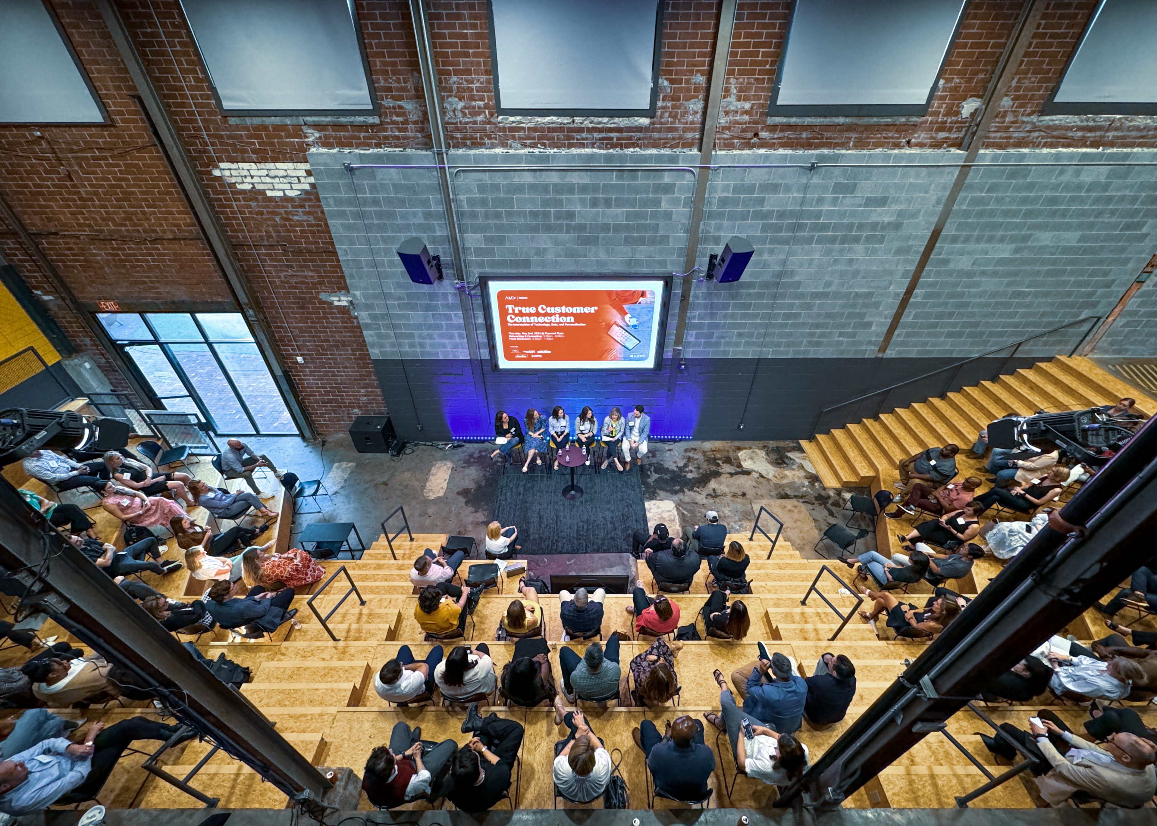 A panel discussion takes place on a stage in a brick-walled venue, with an audience seated on wooden steps.