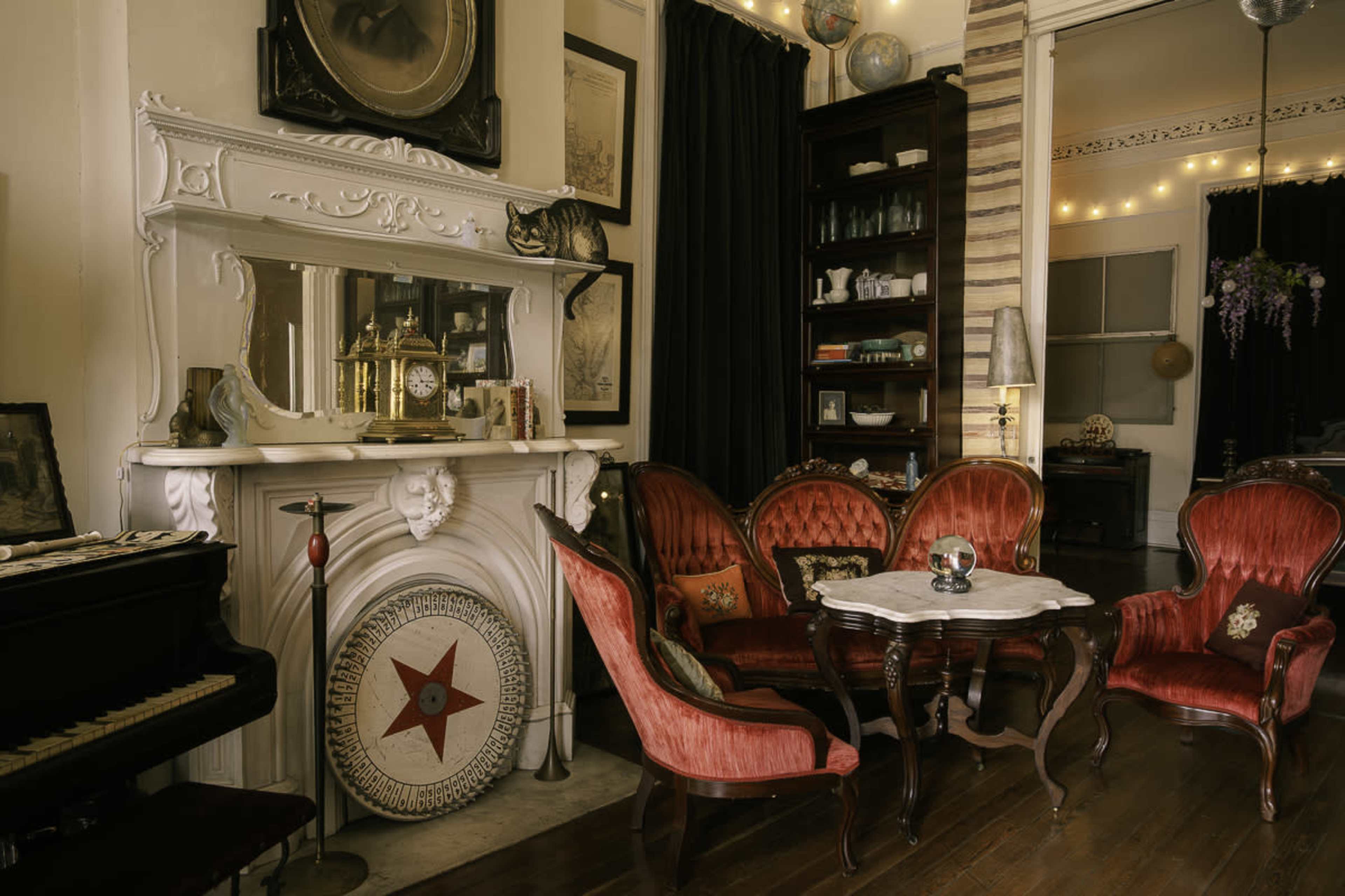 The image depicts a vintage living room featuring ornate red velvet chairs, a white ornate mantelpiece, and a grand piano in a warmly lit setting.